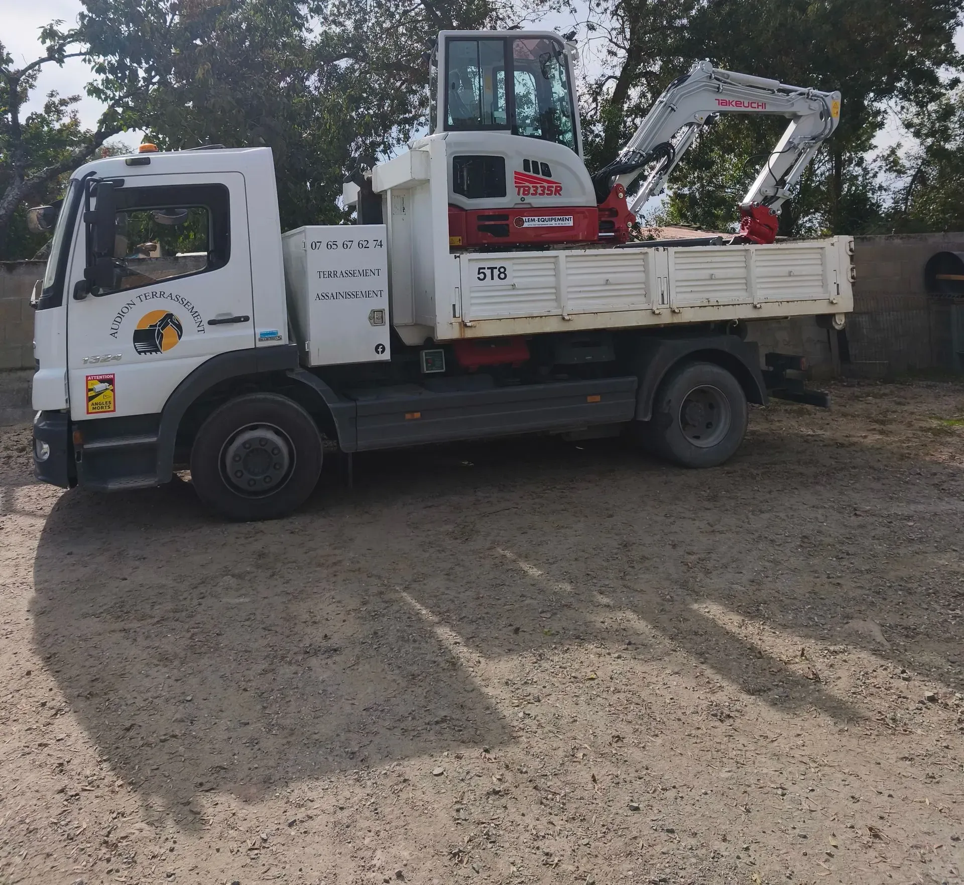 Camion tractant une remorque avec une mini-benne orange sur un terrain gravillonné gris. 