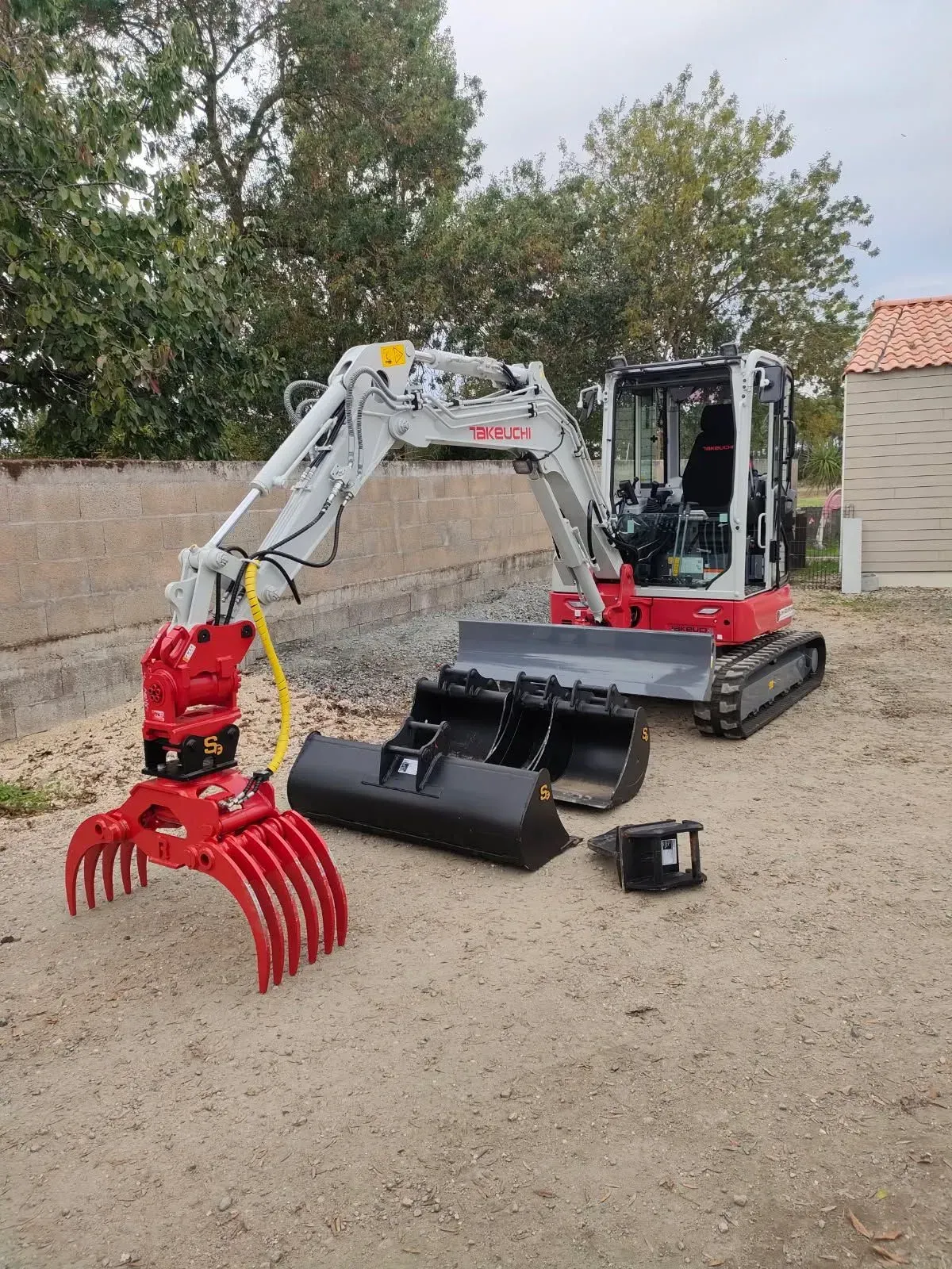 Mini-pelle rouge et blanche avec grappin, godet et attelage dans une cour.