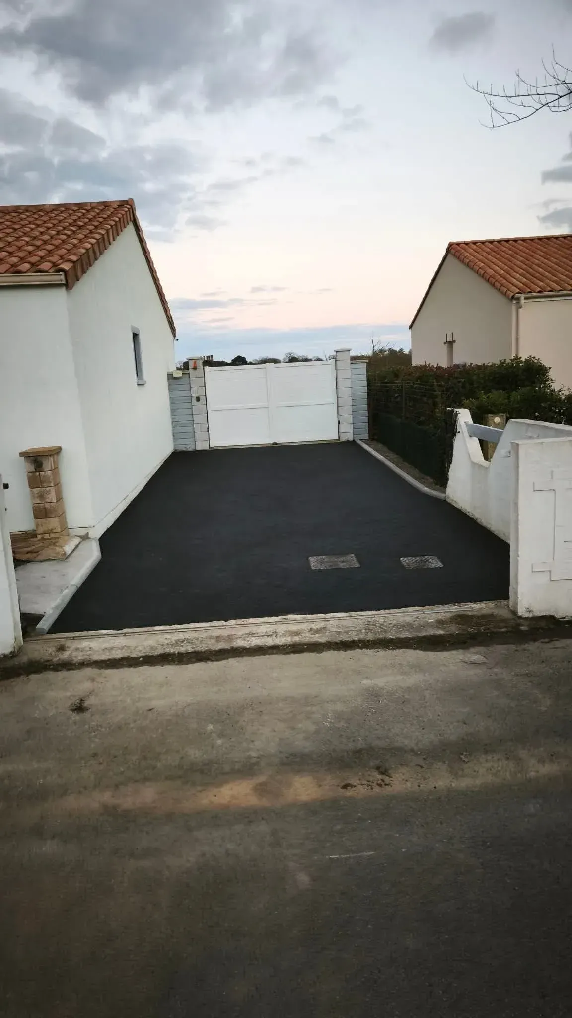Allée asphaltée entre deux maisons blanches aux toits de tuiles rouges. Ciel nuageux en arrière-plan.