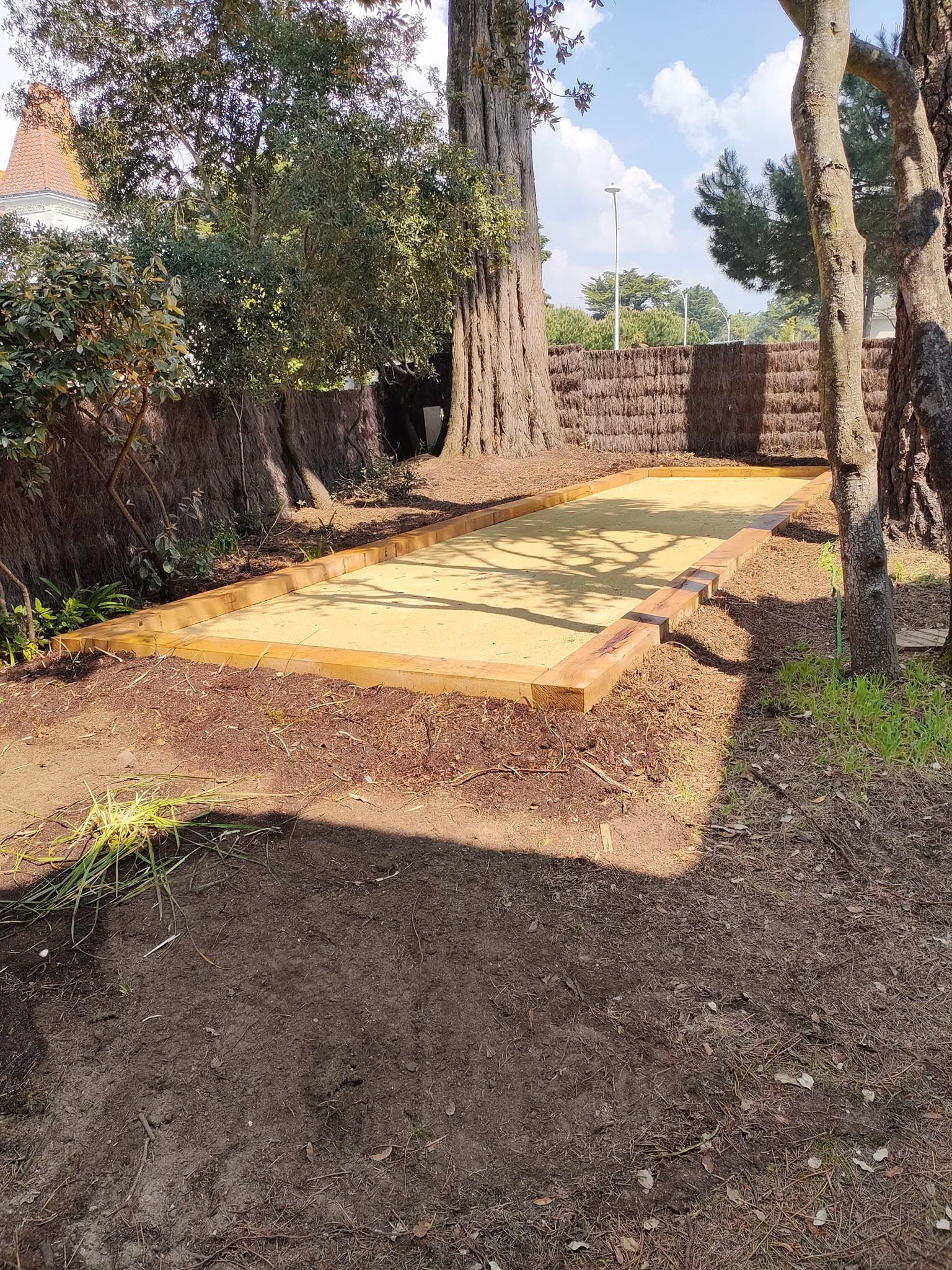 Terrain de pétanque bordé de bois, entouré de terre et d'arbres, cadre extérieur ensoleillé.