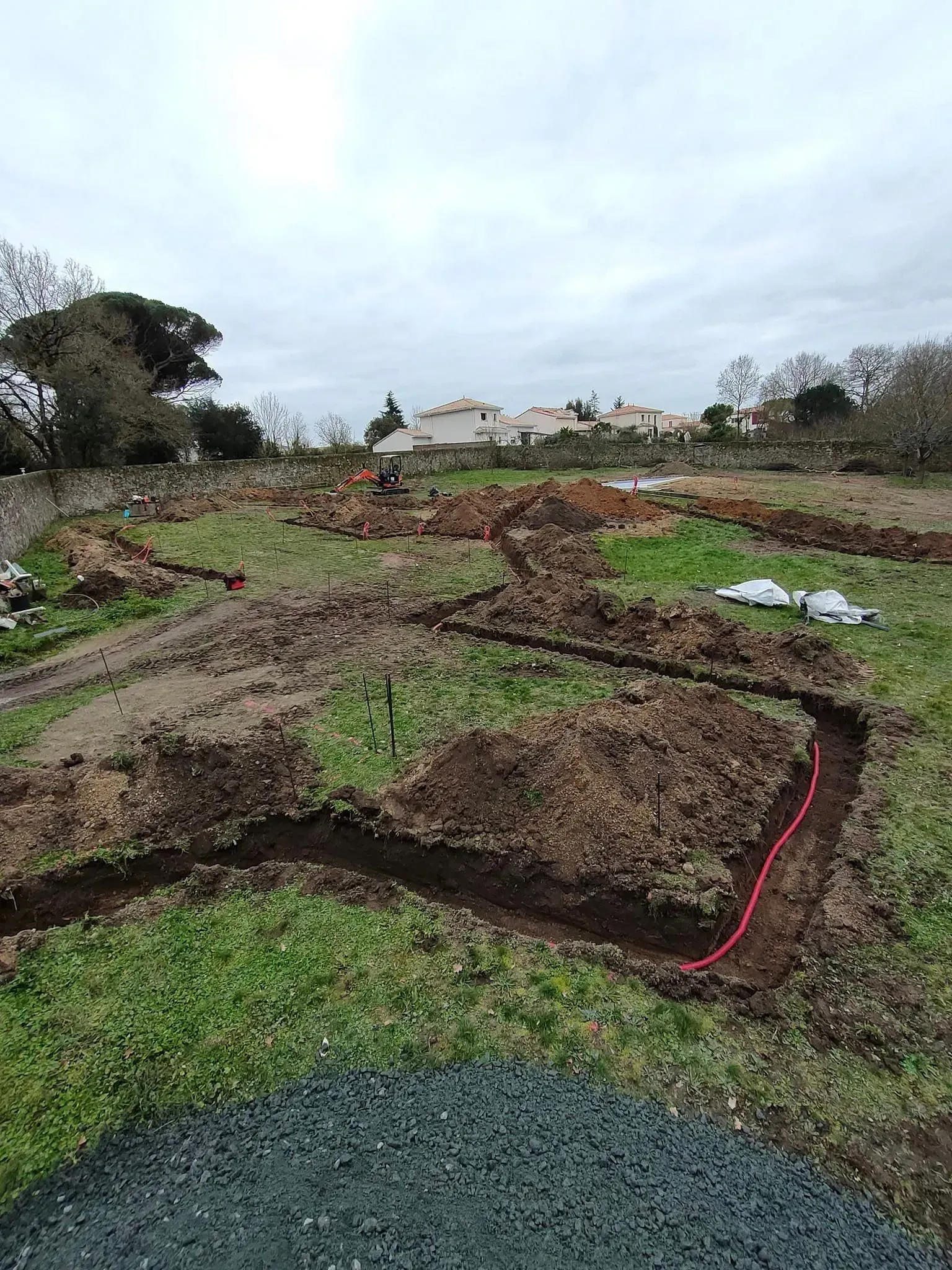 Des tranchées creusées dans un champ herbeux, délimitées en rouge, avec des maisons en arrière-plan sous un ciel nuageux.