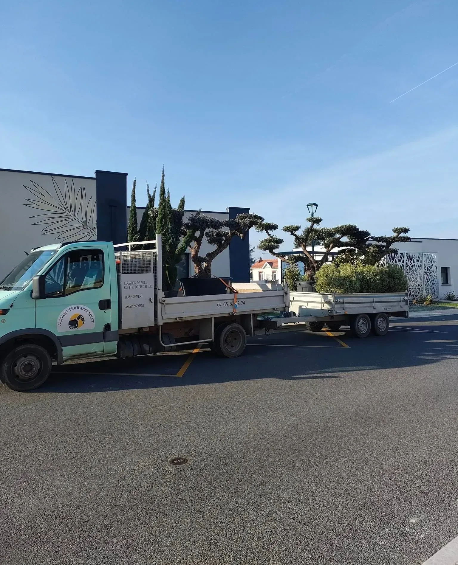 Un camion tirant une remorque chargée d'arbres est stationné devant un bâtiment sous un ciel bleu.