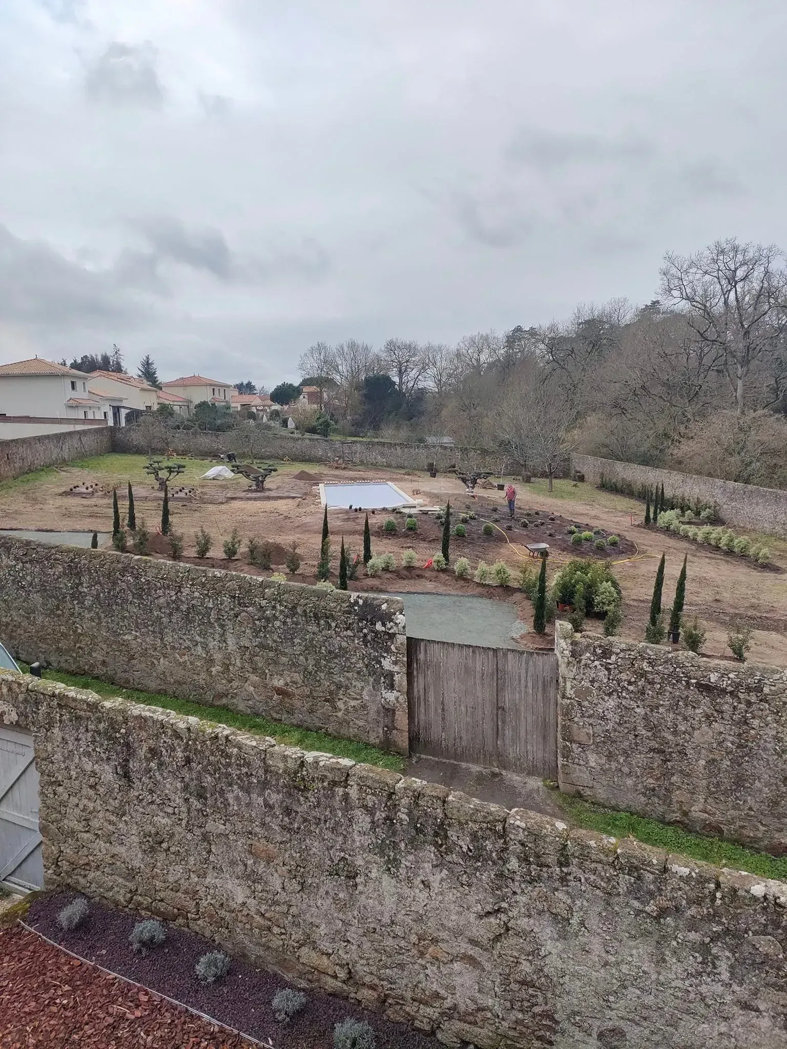Jardin clos de murs en pierre avec portail, arbres et ciel nuageux.