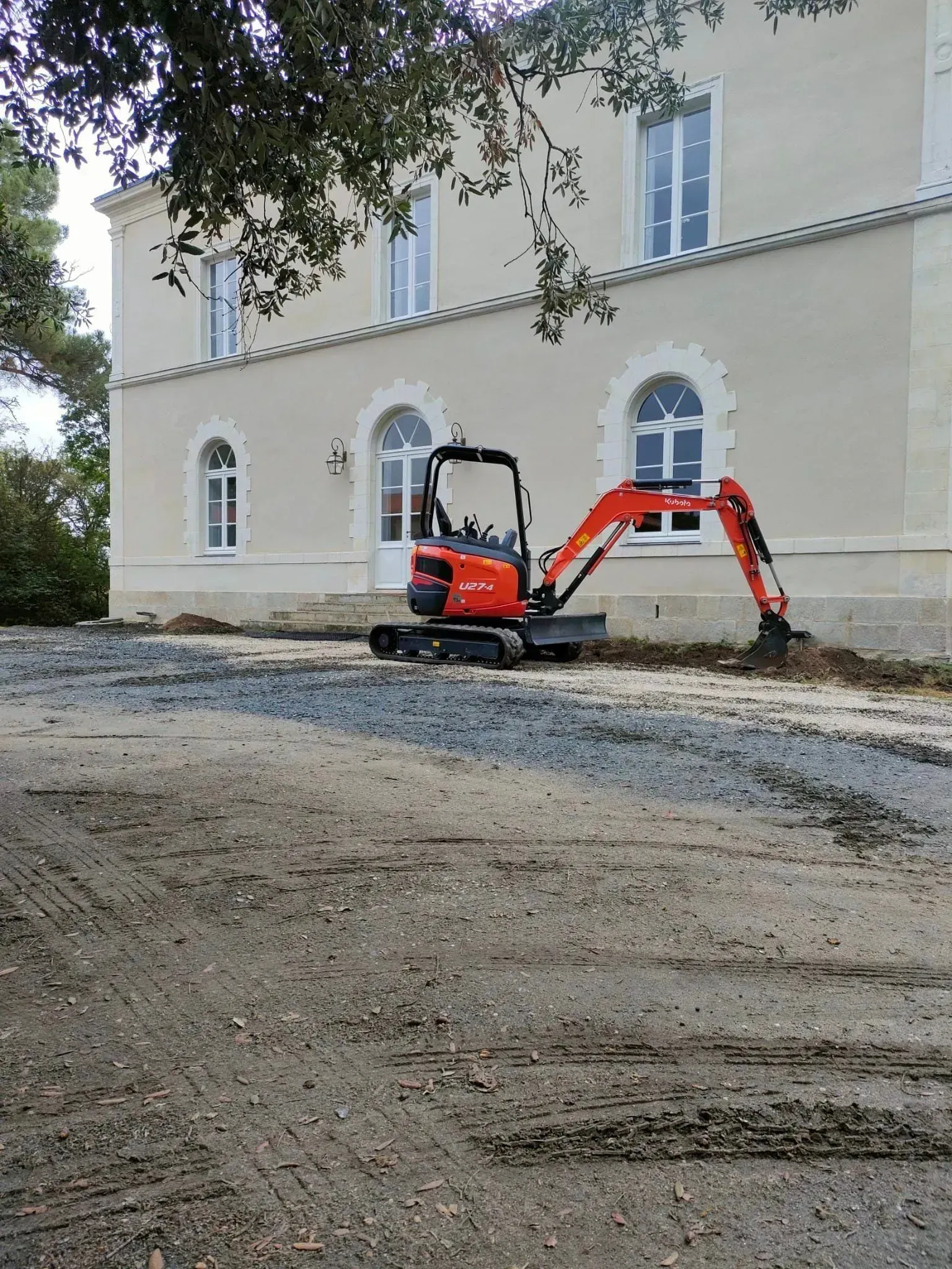 Une pelleteuse orange extrait du gravier à côté d'un bâtiment clair aux fenêtres cintrées.