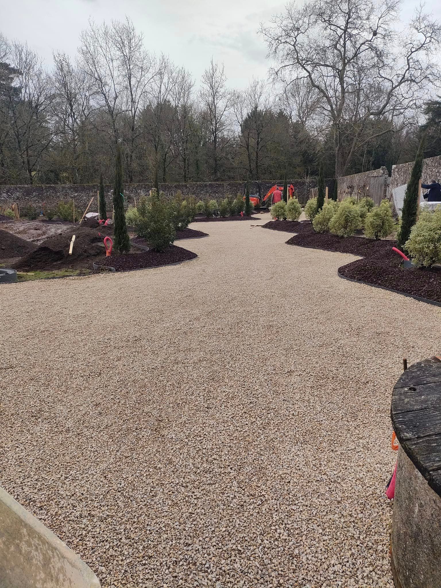 Allée de gravier traversant un jardin avec des arbustes et des arbres nouvellement plantés, sous un ciel nuageux.
