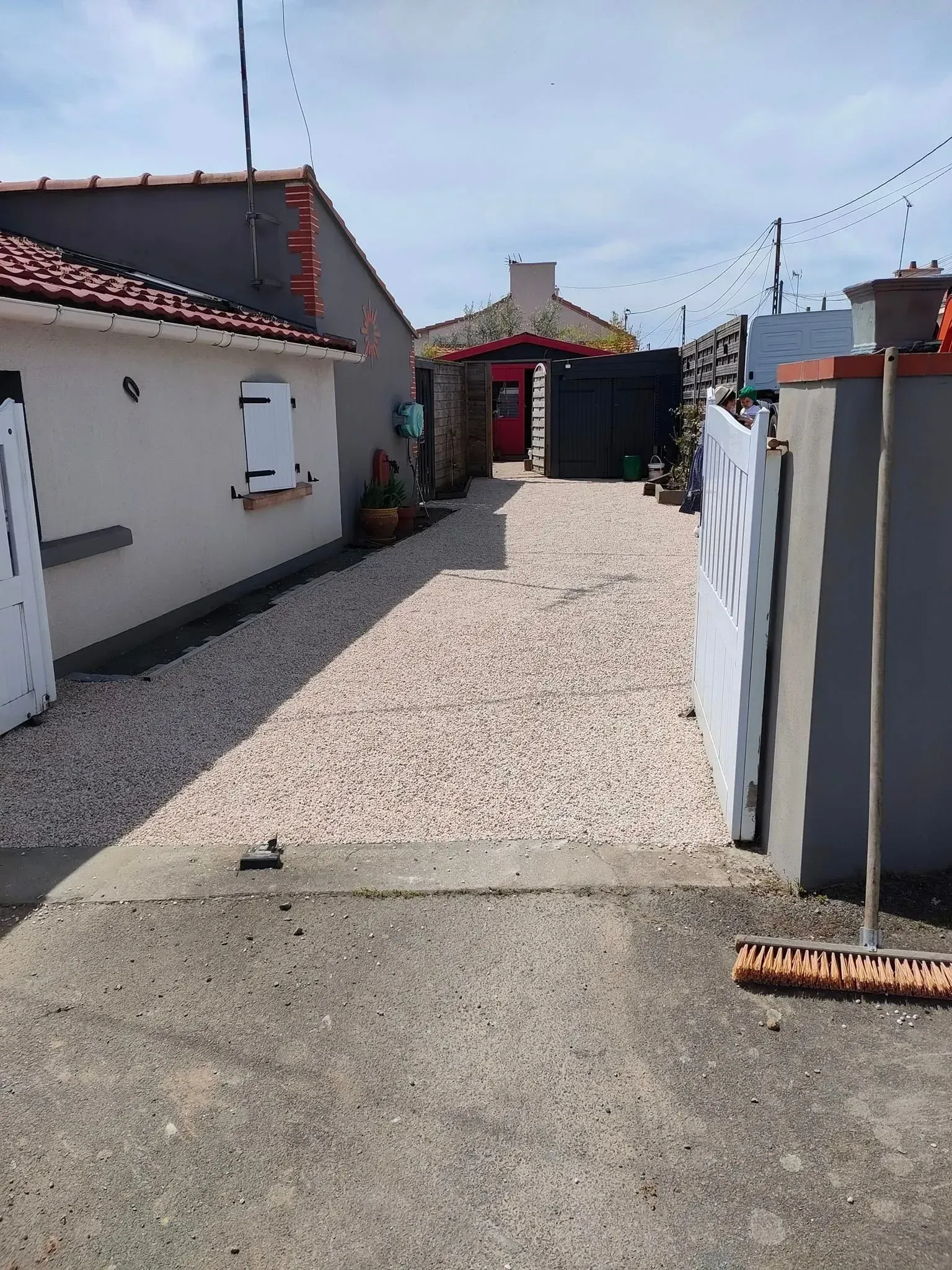 Allée de gravier entre deux bâtiments, avec un mur gris et une clôture blanche à droite, et un bâtiment blanc à gauche.