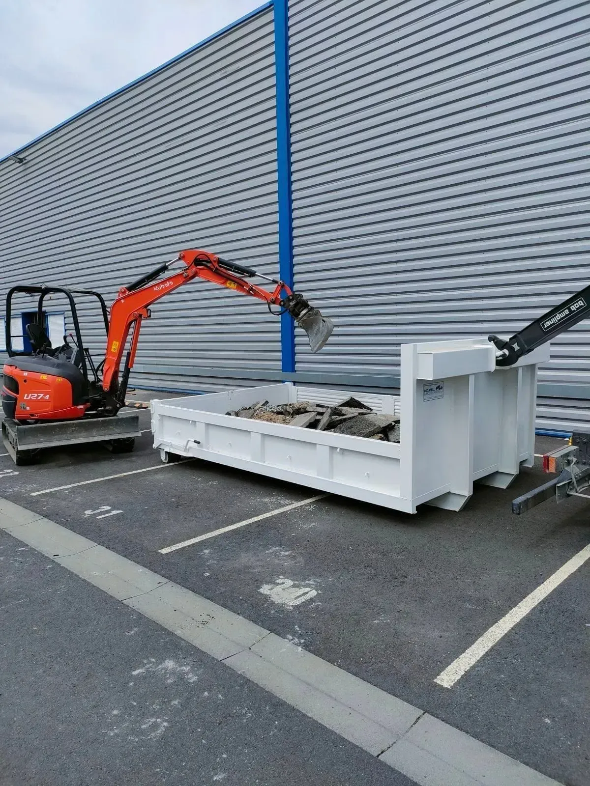Une pelleteuse orange charge des débris dans une benne blanche sur un terrain pavé.