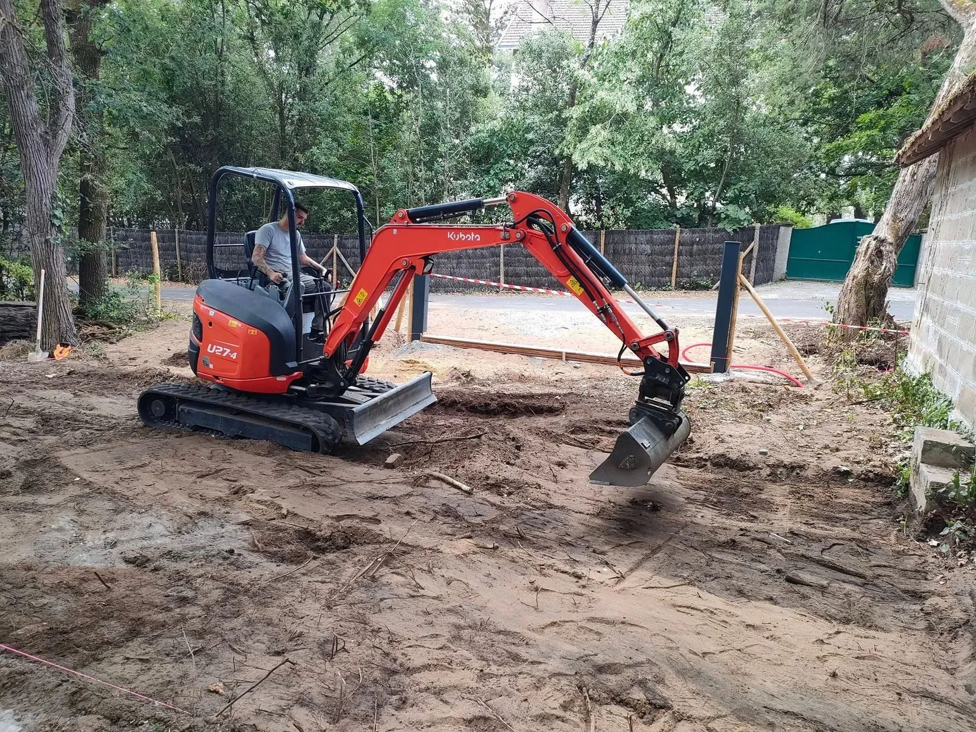 Une petite pelleteuse rouge creuse dans un terrain vague près d'un bâtiment et d'arbres.