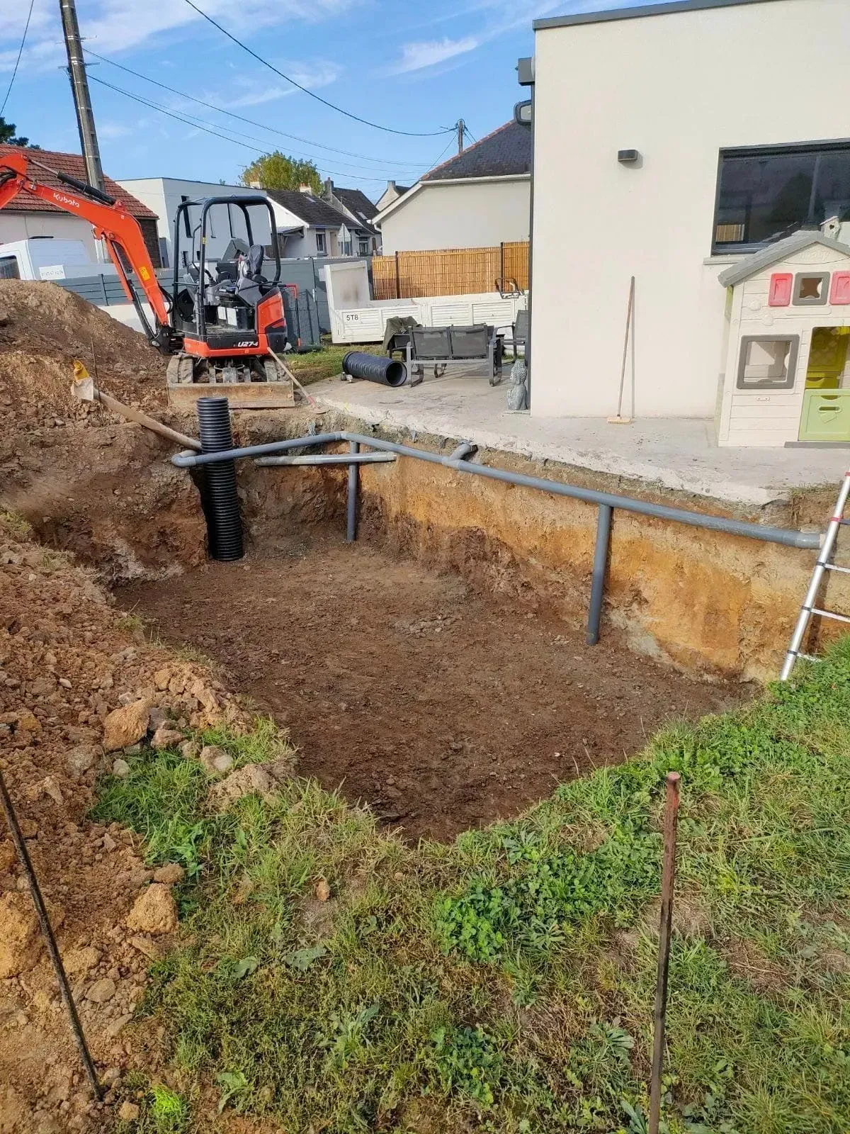 Chantier de fouilles avec une mini-pelle. Une tranchée est ouverte près d'un bâtiment et d'une petite maisonnette de jeux.