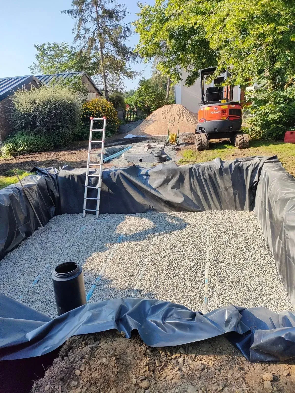 Construction d'un bassin rectangulaire dans un jardin, rempli de gravier, bâche noire, échelle. Mini-pelle en arrière-plan.