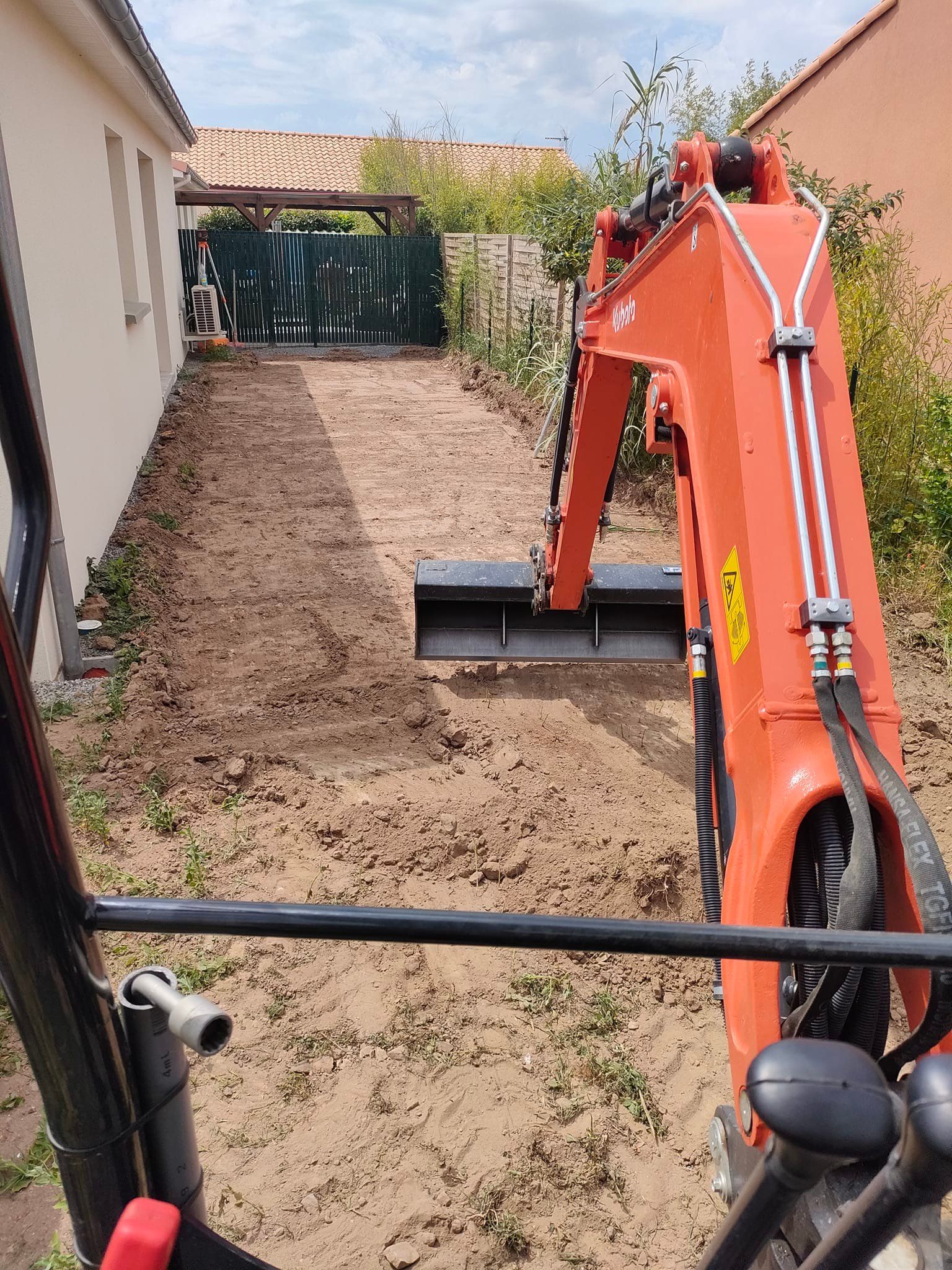 Une pelleteuse orange creuse dans une cour étroite en terre battue, entre un bâtiment et une clôture.