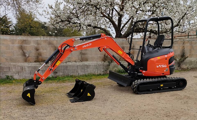 Une mini-pelle Kubota orange avec deux godets, posée au sol devant un muret en pierre et un arbre en fleurs.
