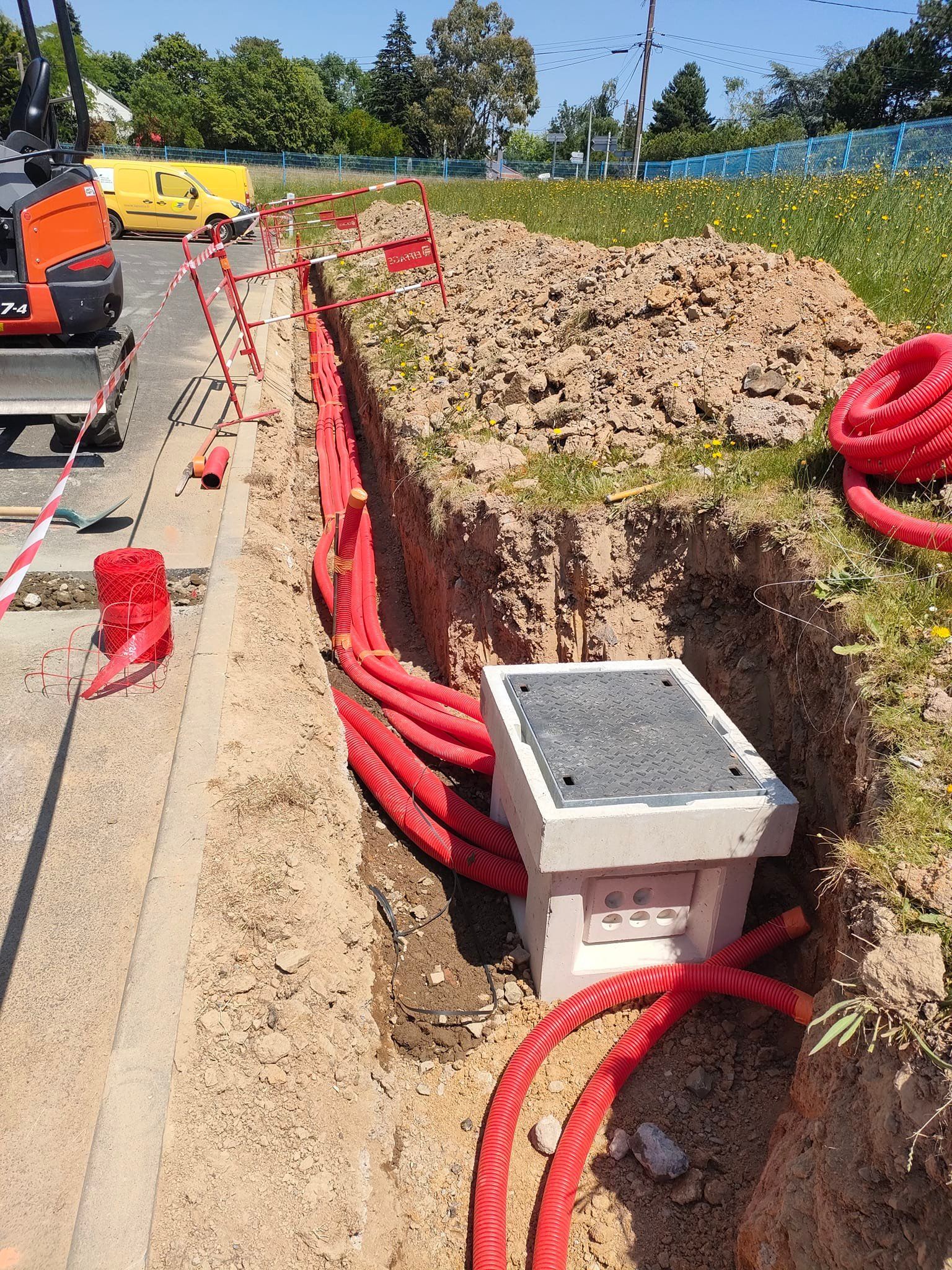 Tranchée avec conduits rouges, boîte électrique et petite pelle mécanique. Chantier par une journée ensoleillée.