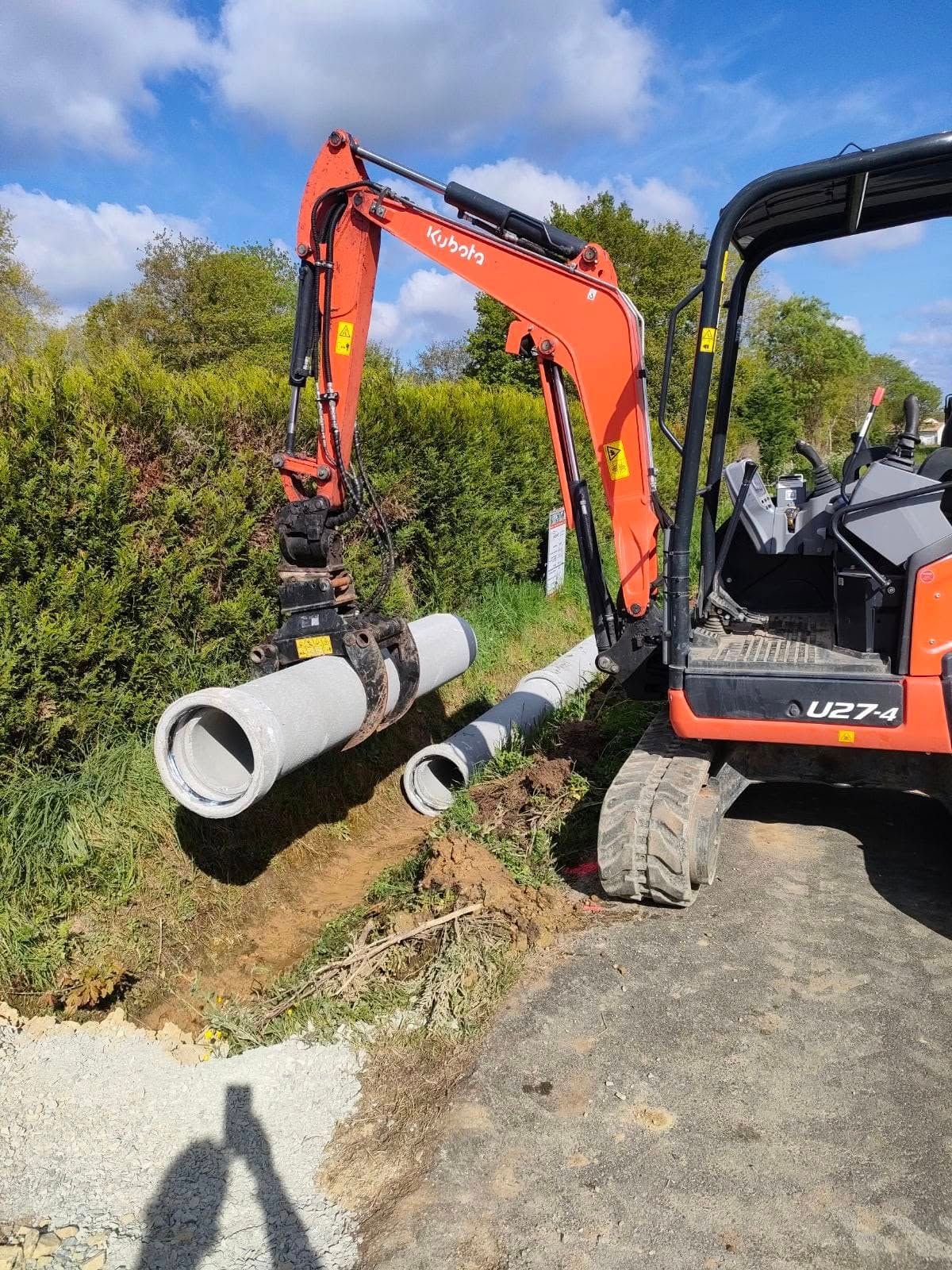 Une pelleteuse orange pose des tuyaux de drainage en béton au bord d'une route. 