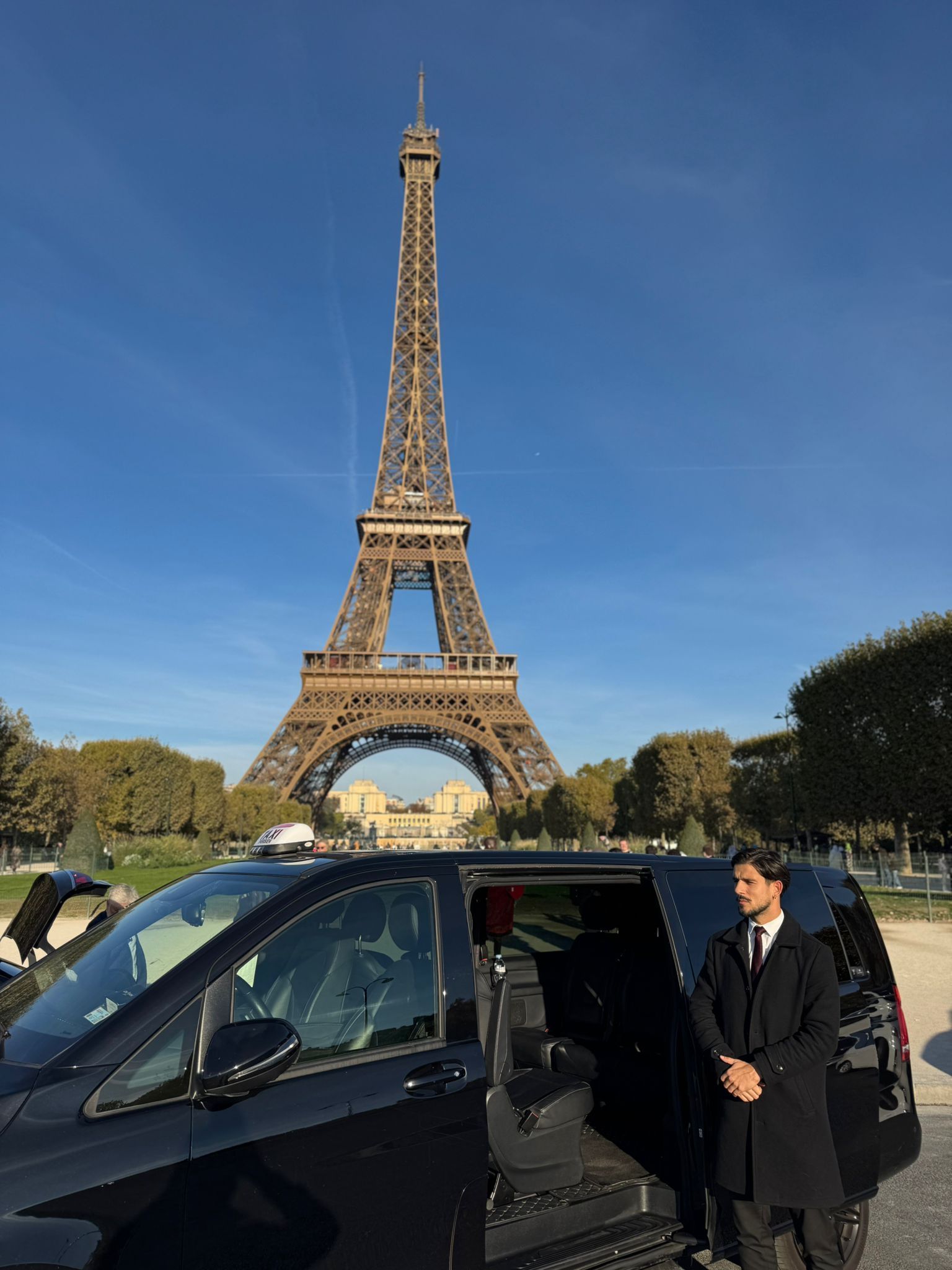 Un homme en costume et lunettes de soleil se tient près d'une voiture noire, les bras croisés.