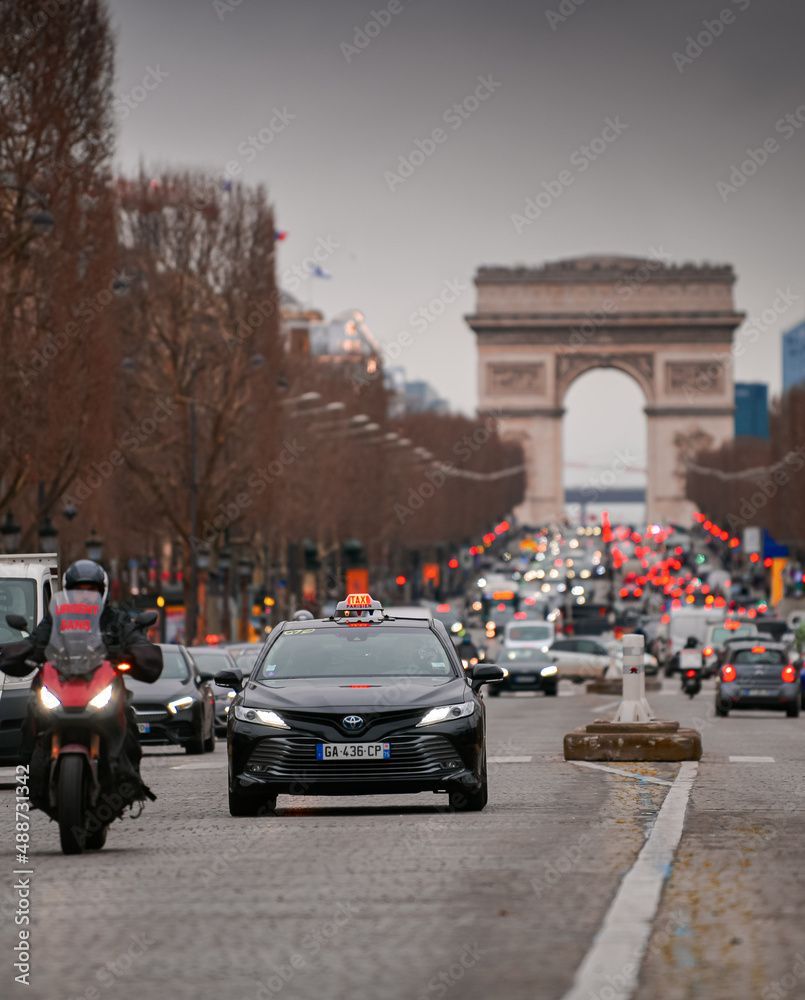 Des voitures et une moto circulant dans une large rue de Paris avec l'Arc de Triomphe en arrière-plan.