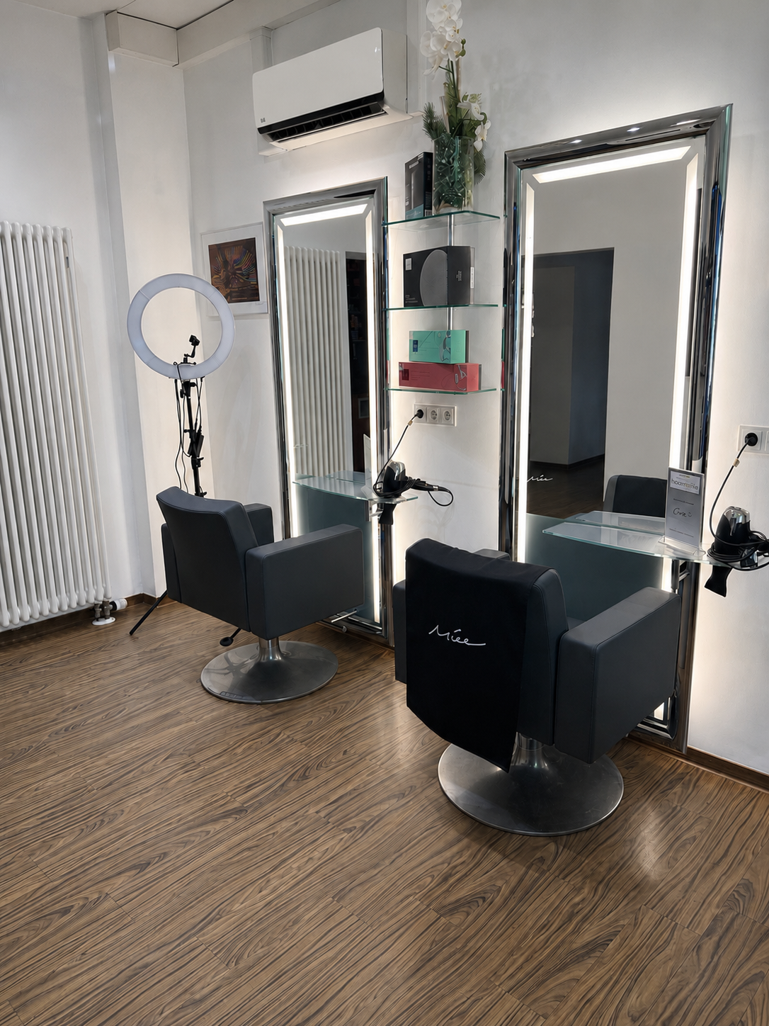 Saloneinrichtung mit zwei schwarzen Friseurstühlen, Spiegeln, Regalen und einem Ringlicht auf einem Holzboden.