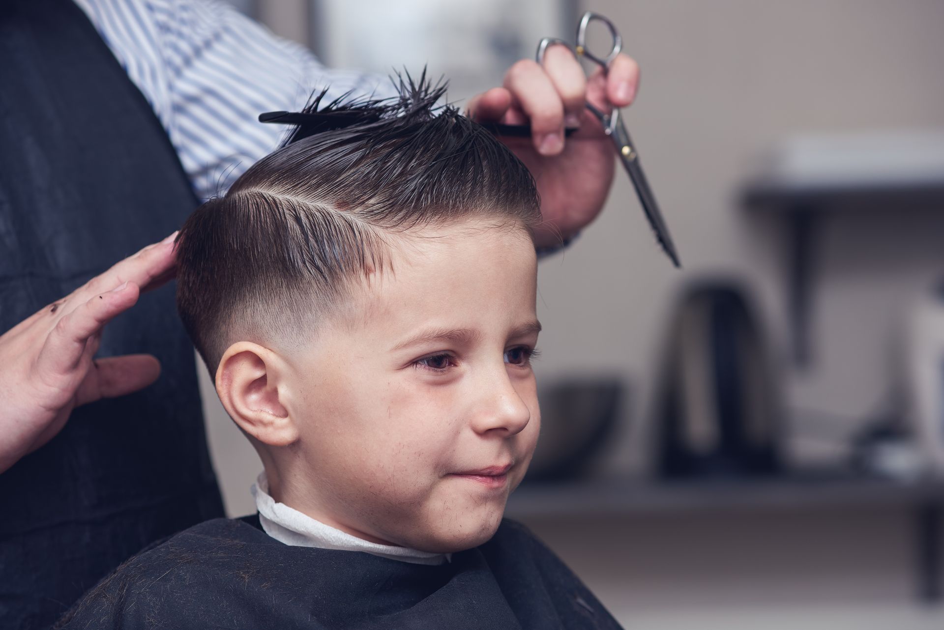 Ein Kind, dem im Friseursalon mit Schere und Kamm die Haare geschnitten werden, lächelt leicht.