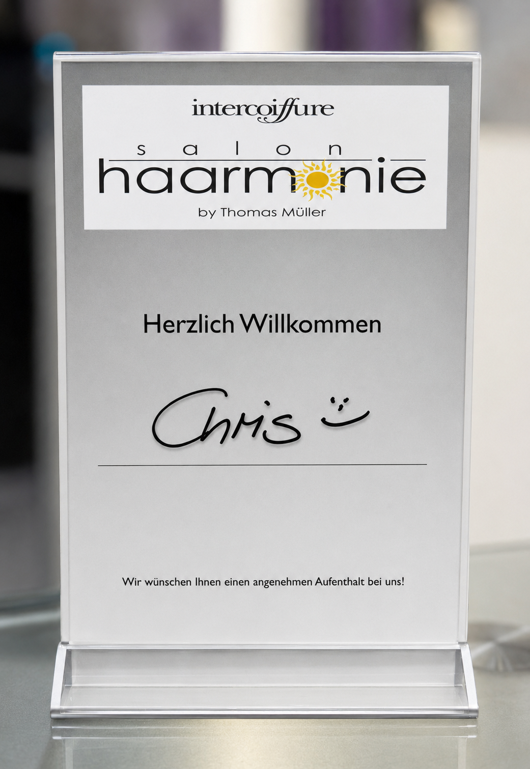 Schild auf einem durchsichtigen Ständer mit „härmonie“, „Herzlich Willkommen“ und darunter handgeschrieben „Chris“.