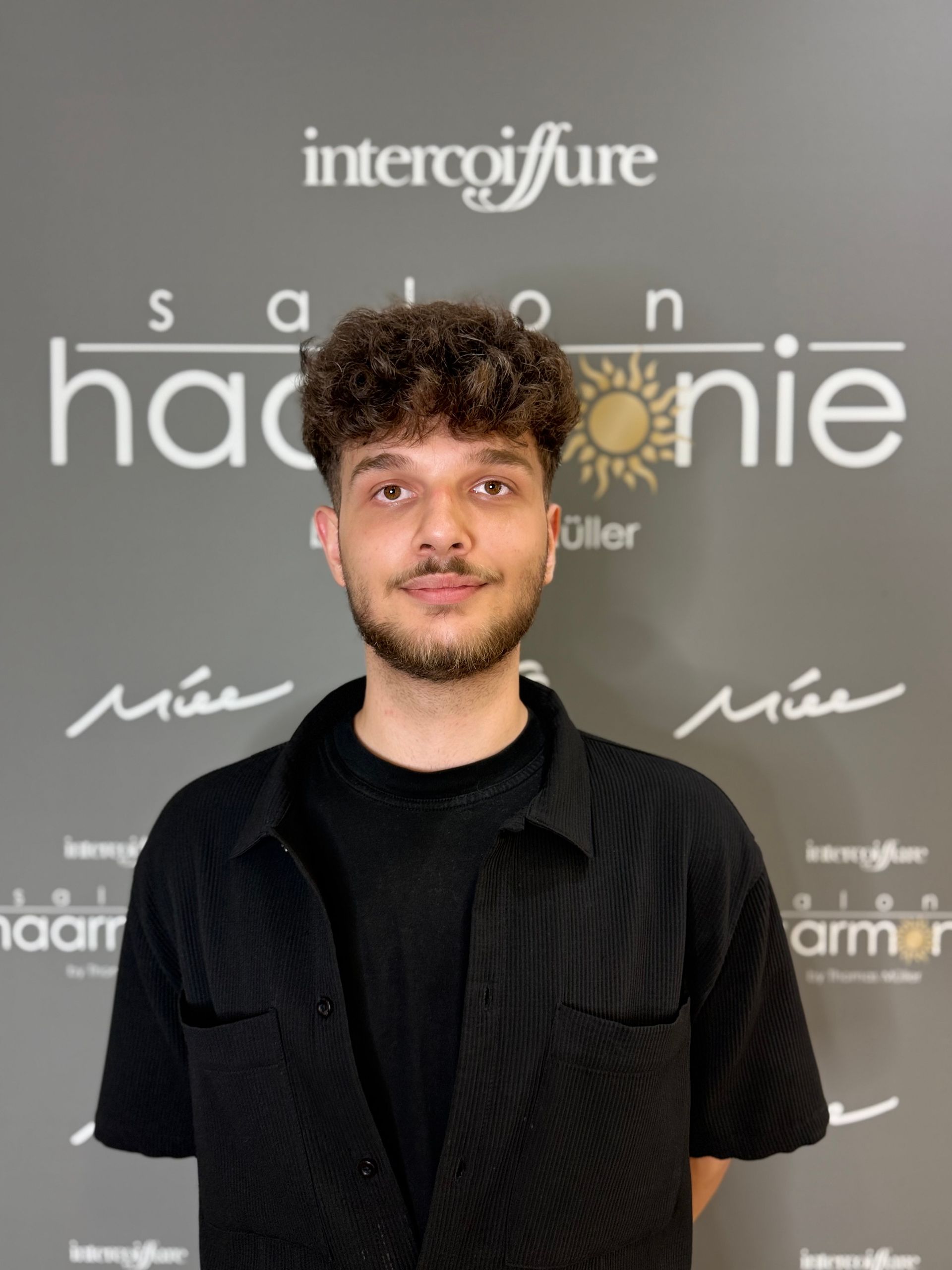 Person mit lockigem Haar in einem schwarzen Hemd vor einer grauen Veranstaltungswand mit weißen Logos.