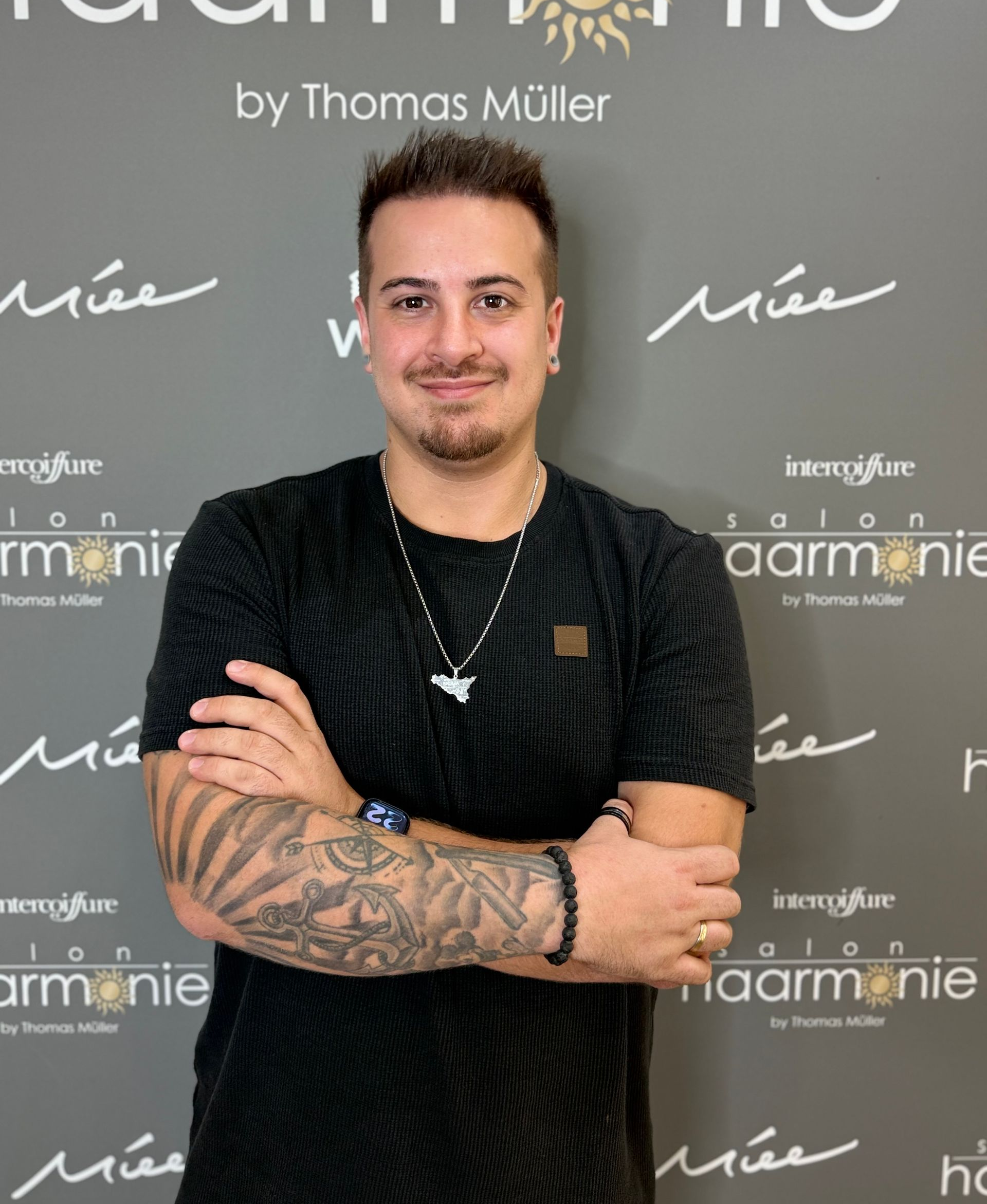 Mann mit verschränkten Armen vor einem schwarzen Hintergrund mit Markenlogo, in schwarzem Hemd und mit Halskette.