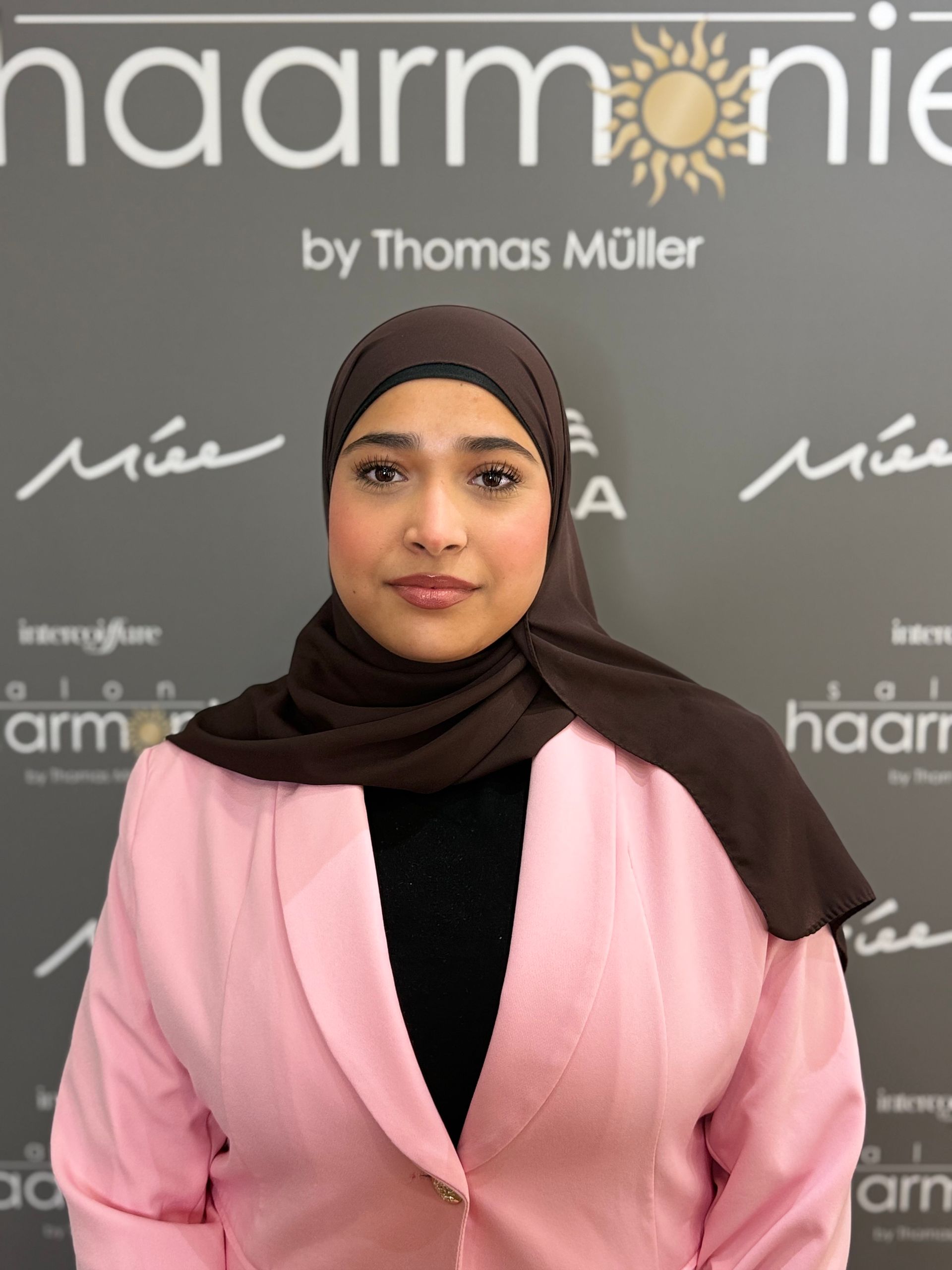 Frau mit schwarzem Hijab und rosafarbenem Blazer vor einer Haarmonie by Thomas Müller-Kulisse posierend