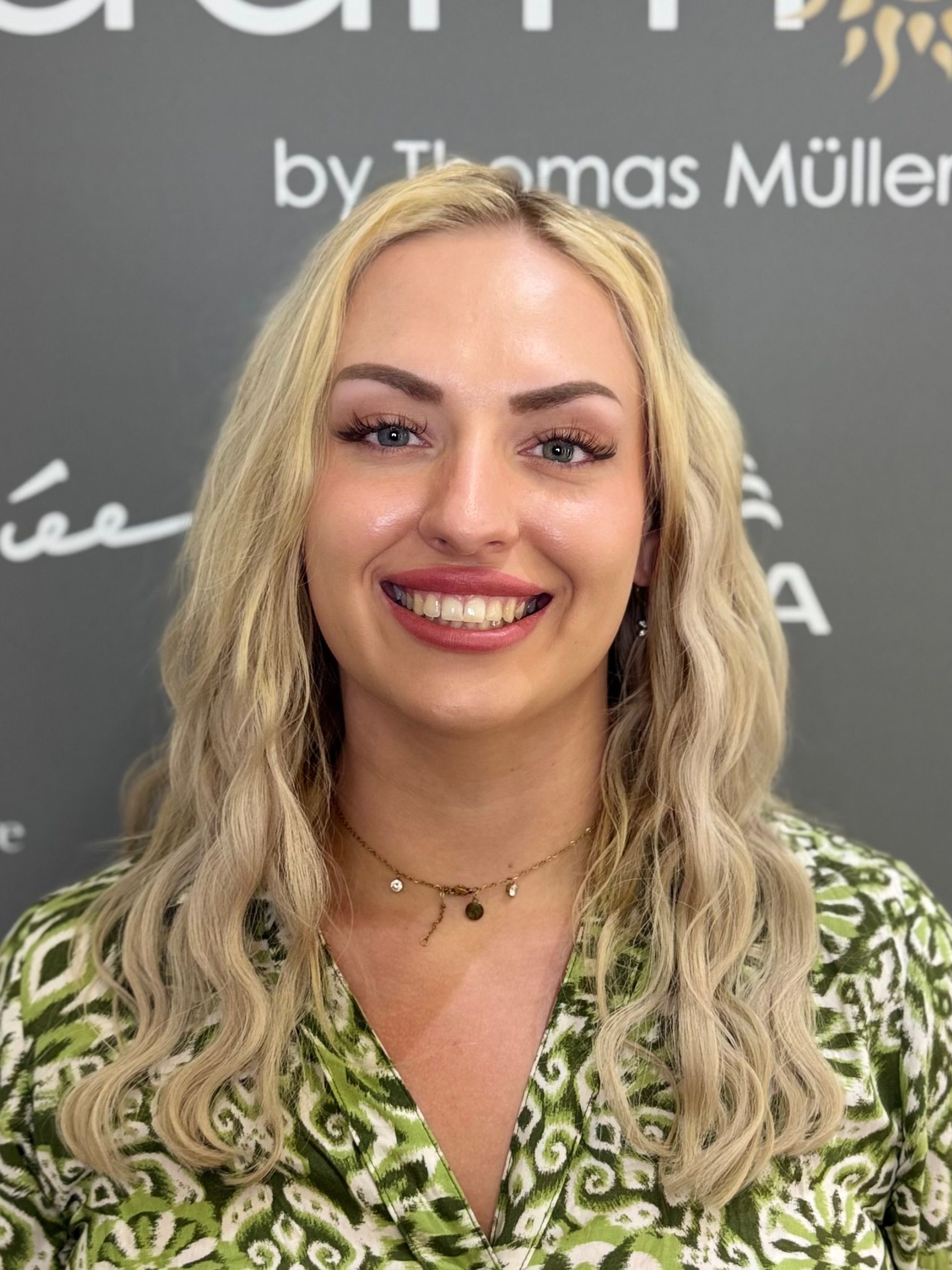 Lächelnde blonde Frau in einer grünen gemusterten Bluse vor einem dunklen Hintergrund mit Markenlogo poseierend