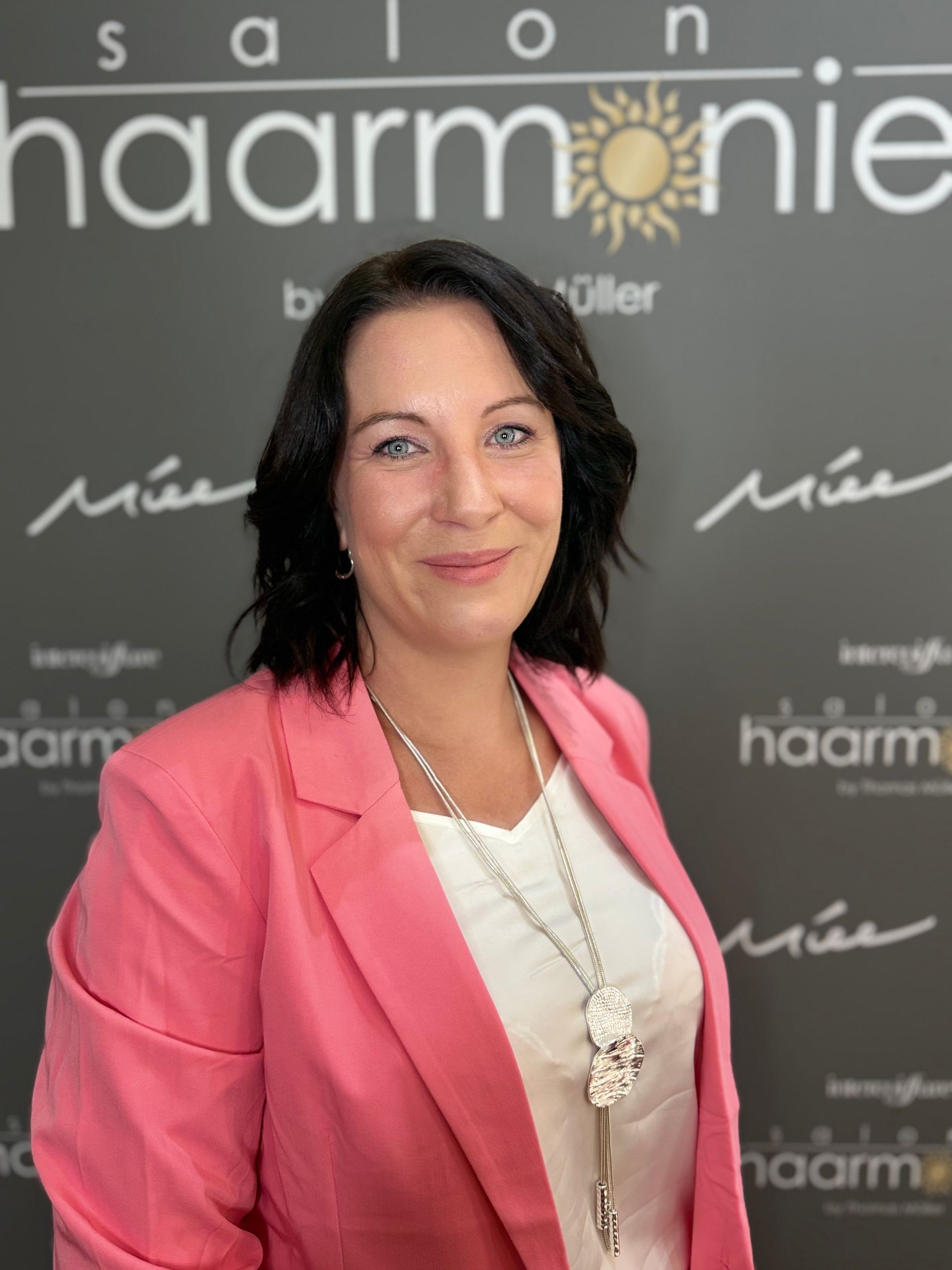 Frau in einem rosa Blazer lächelt vor einer Haarmonie-Kulisse und trägt eine lange Halskette.