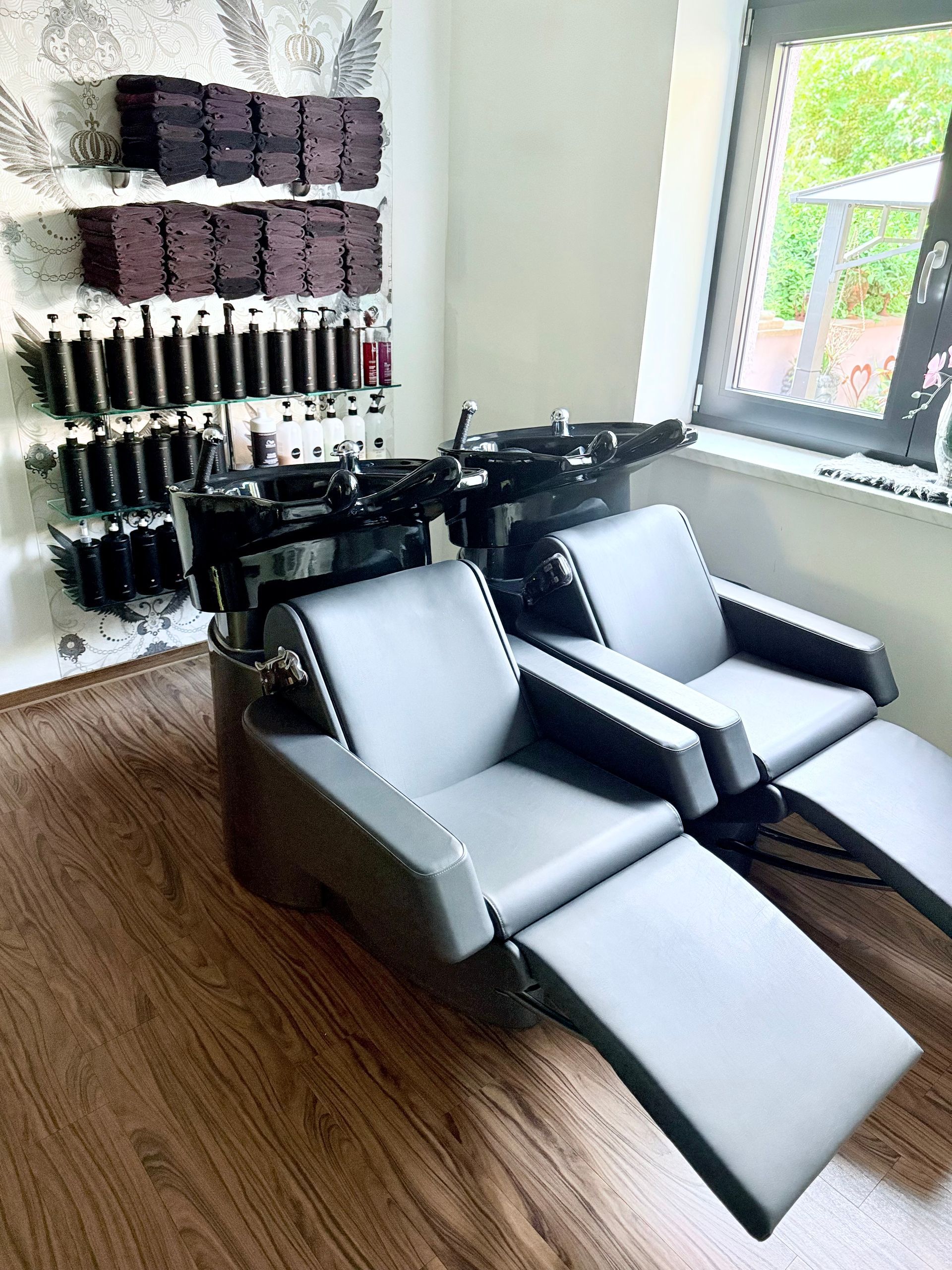 Friseurstühle mit Waschbecken vor einem Fenster, an der Wand Regale mit Haarpflegeprodukten
