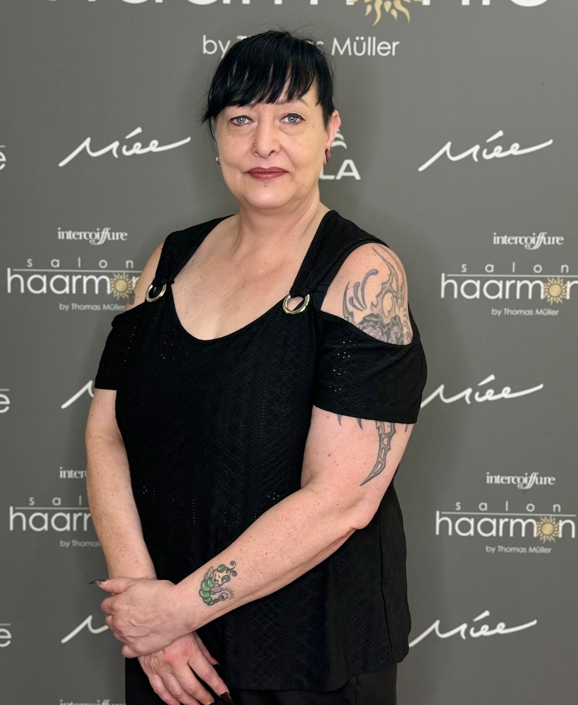 Frau in schwarzem Kleid posiert vor einer gebrandeten Kulisse, Tattoos an den Armen sichtbar.