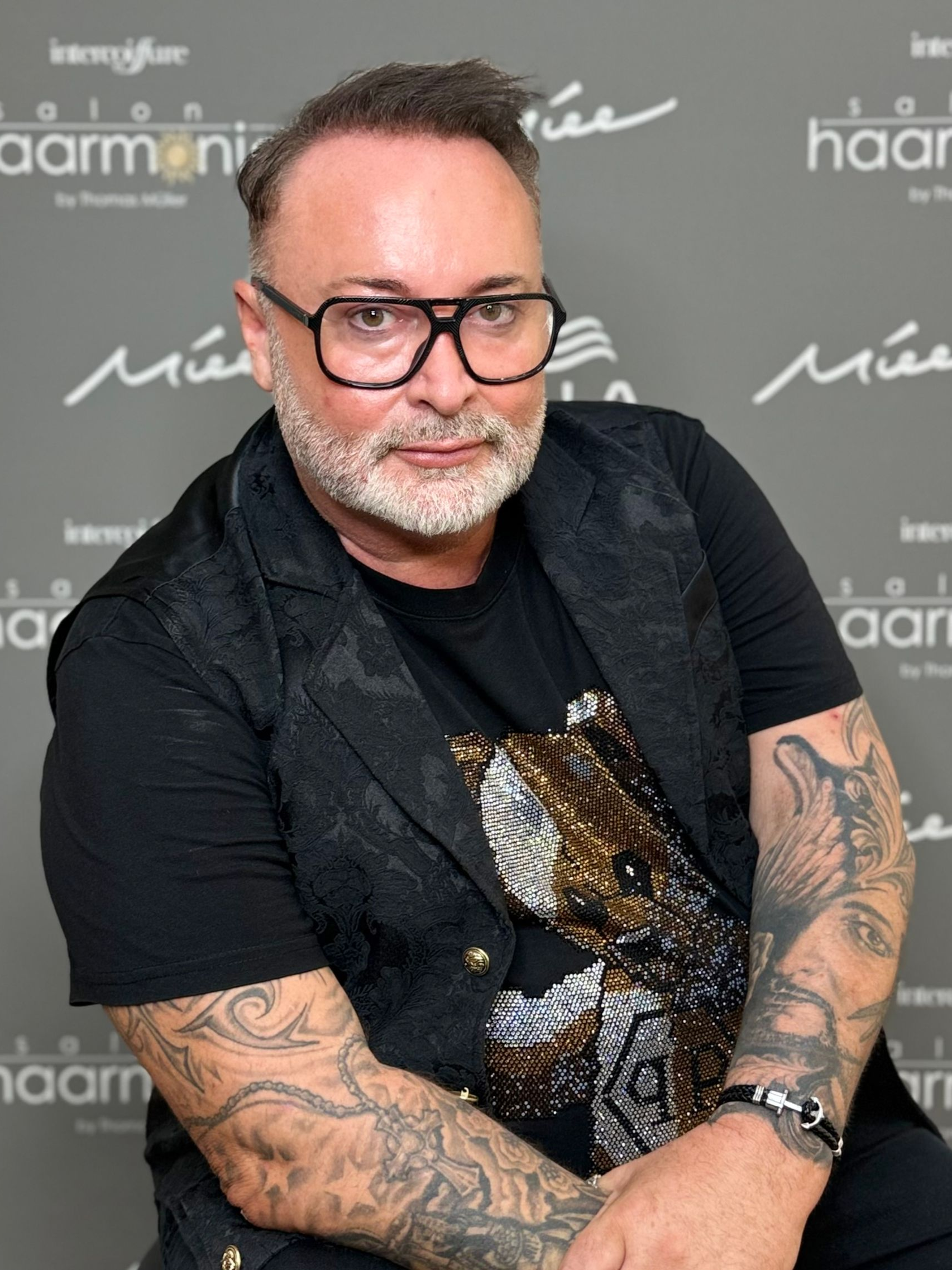 Ein Mann mit Brille und tätowierten Armen posiert in einem schwarzen Hemd vor einer Sponsorenwand