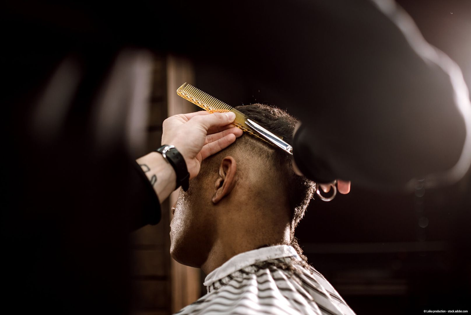 Ein Friseur stutzt einem Kunden in einem schwach beleuchteten Friseursalon mit Haarschneidemaschine und Kamm die Haare.