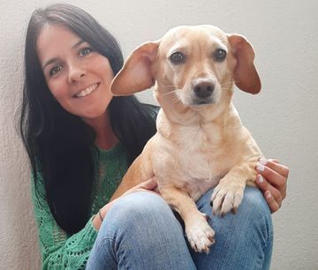 Una mujer de cabello largo y oscuro, sonriente, sostiene un perro bronceado en su regazo; ambos miran a la cámara.