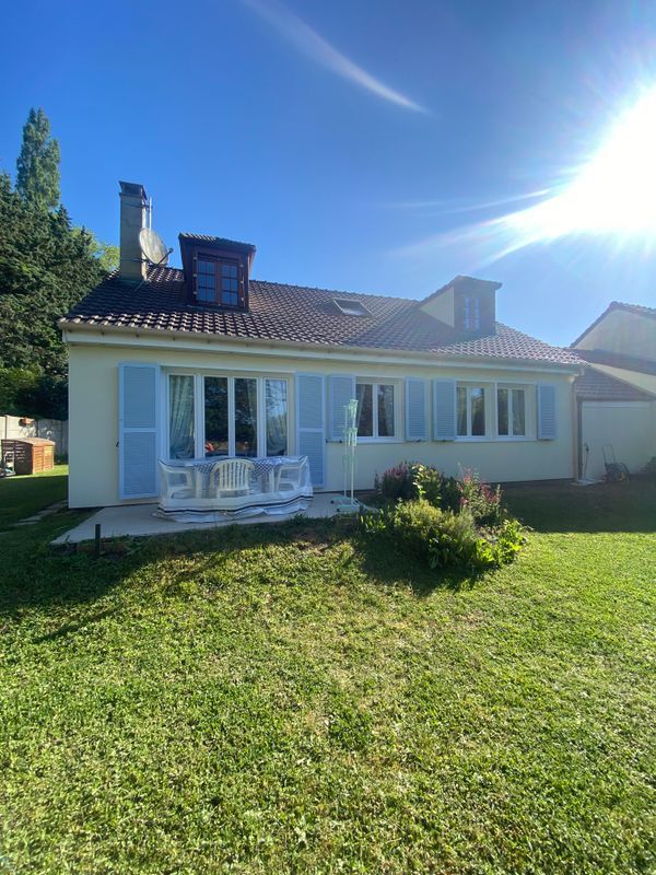Maison blanche de plain-pied avec volets bleus et toit de tuiles, sur une pelouse ensoleillée, avec un éclat de soleil au-dessus.