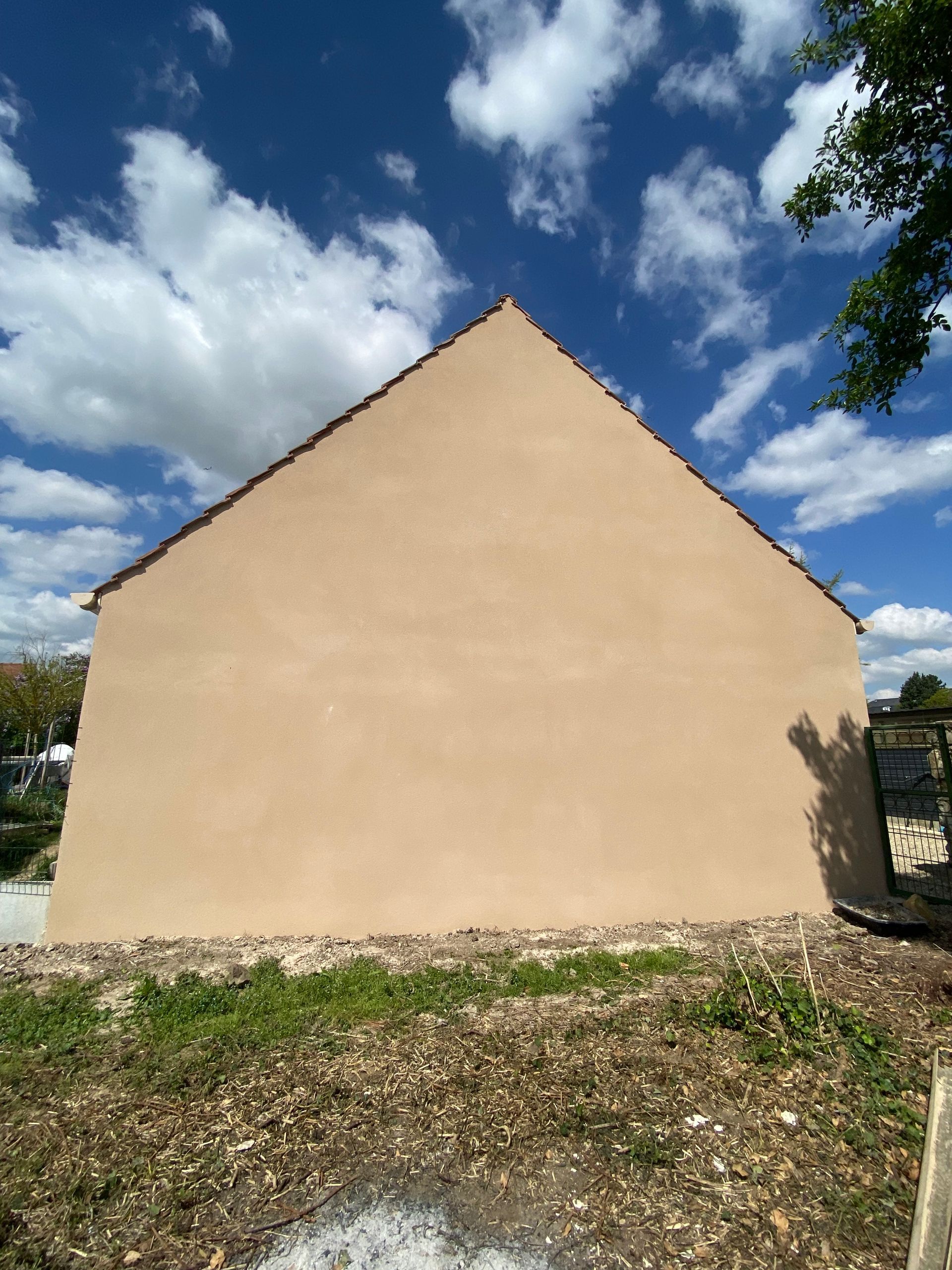 Mur et toiture beiges d'une maison sous un ciel bleu nuageux, vus depuis une cour de gravier.