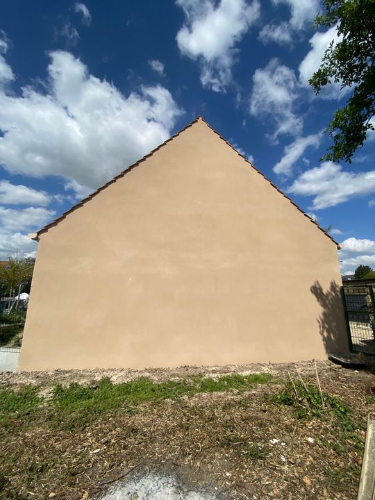 Mur et toiture beiges d'une maison sous un ciel bleu nuageux, vus depuis une cour de gravier.