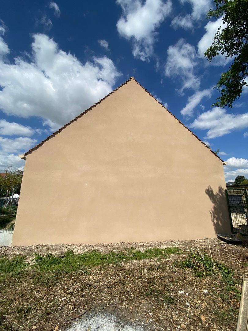 Mur et toiture beiges d'une maison sous un ciel bleu nuageux, vus depuis une cour de gravier.