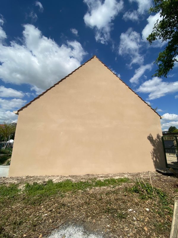 Pignon d'une maison en stuc beige sous un ciel bleu clair nuageux, entouré d'herbe et de terre.