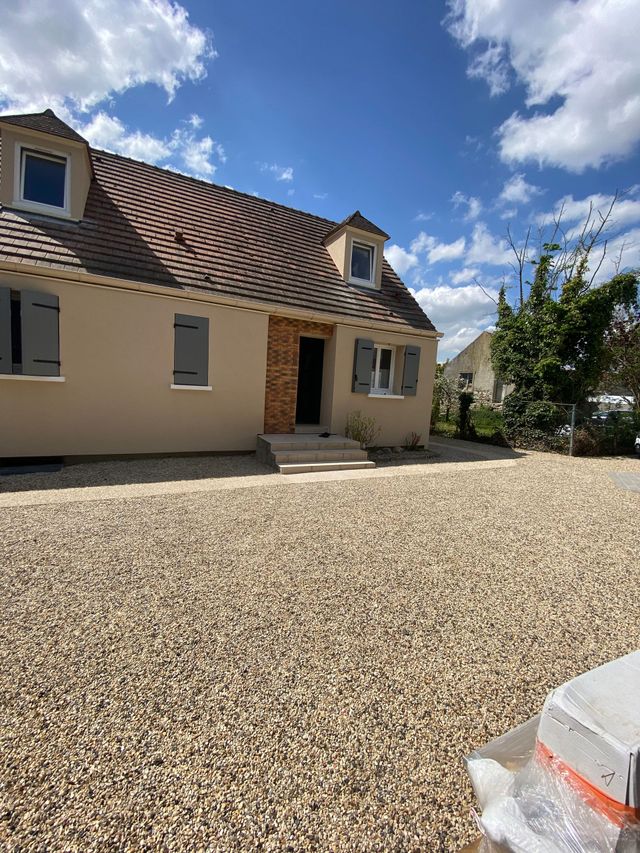 Vue de face d'une maison beige avec un toit de tuiles et une allée de gravier sous un ciel bleu