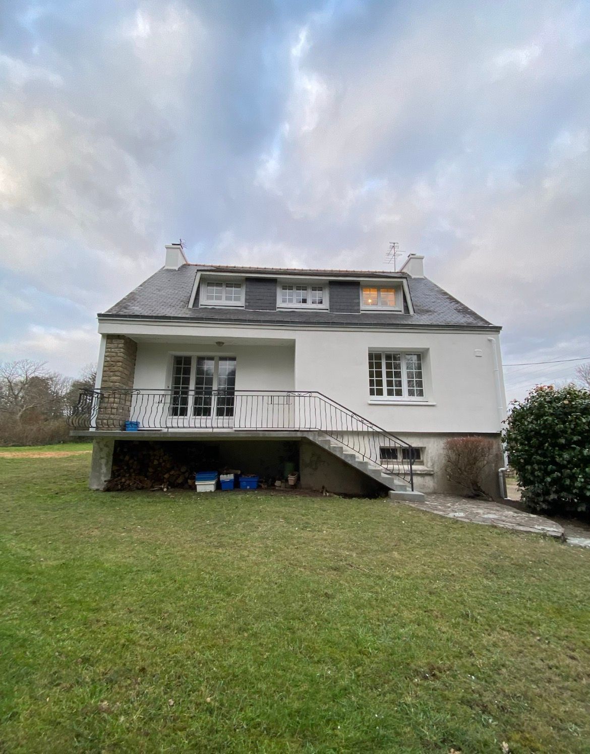 Maison blanche à deux étages avec lucarnes et porche, située sur une pelouse sous un ciel nuageux.