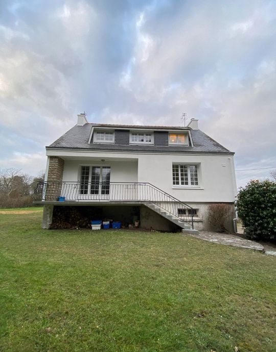 Maison blanche à deux étages avec lucarnes et porche, située sur une pelouse sous un ciel nuageux.