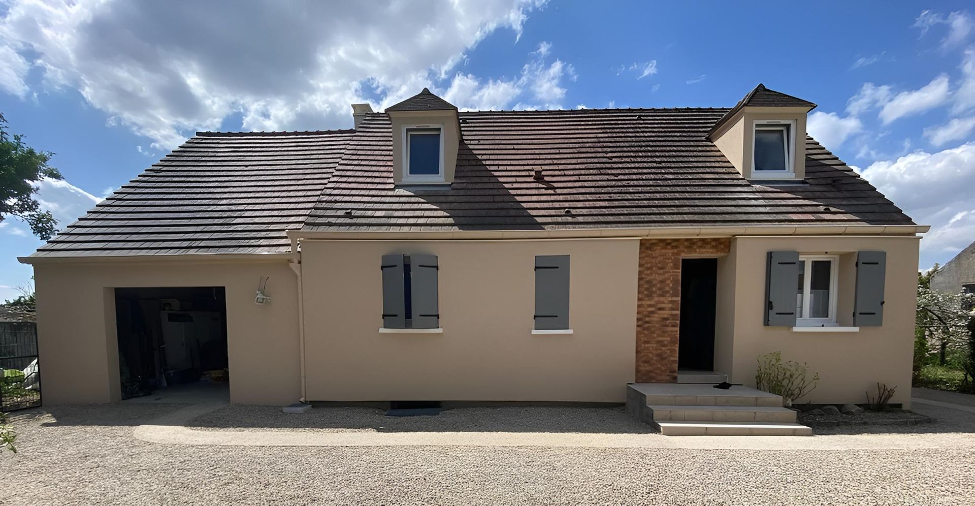 Maison beige avec un toit de tuiles, des lucarnes et un garage ouvert sous un ciel bleu
