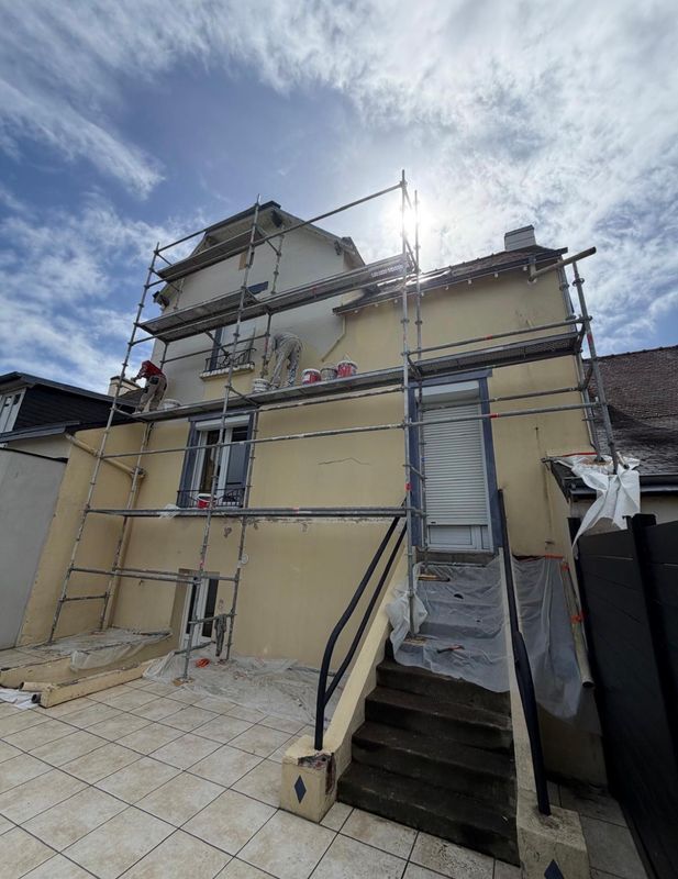 Échafaudages autour d'une maison beige en rénovation, avec des escaliers et un ciel nuageux au-dessus.