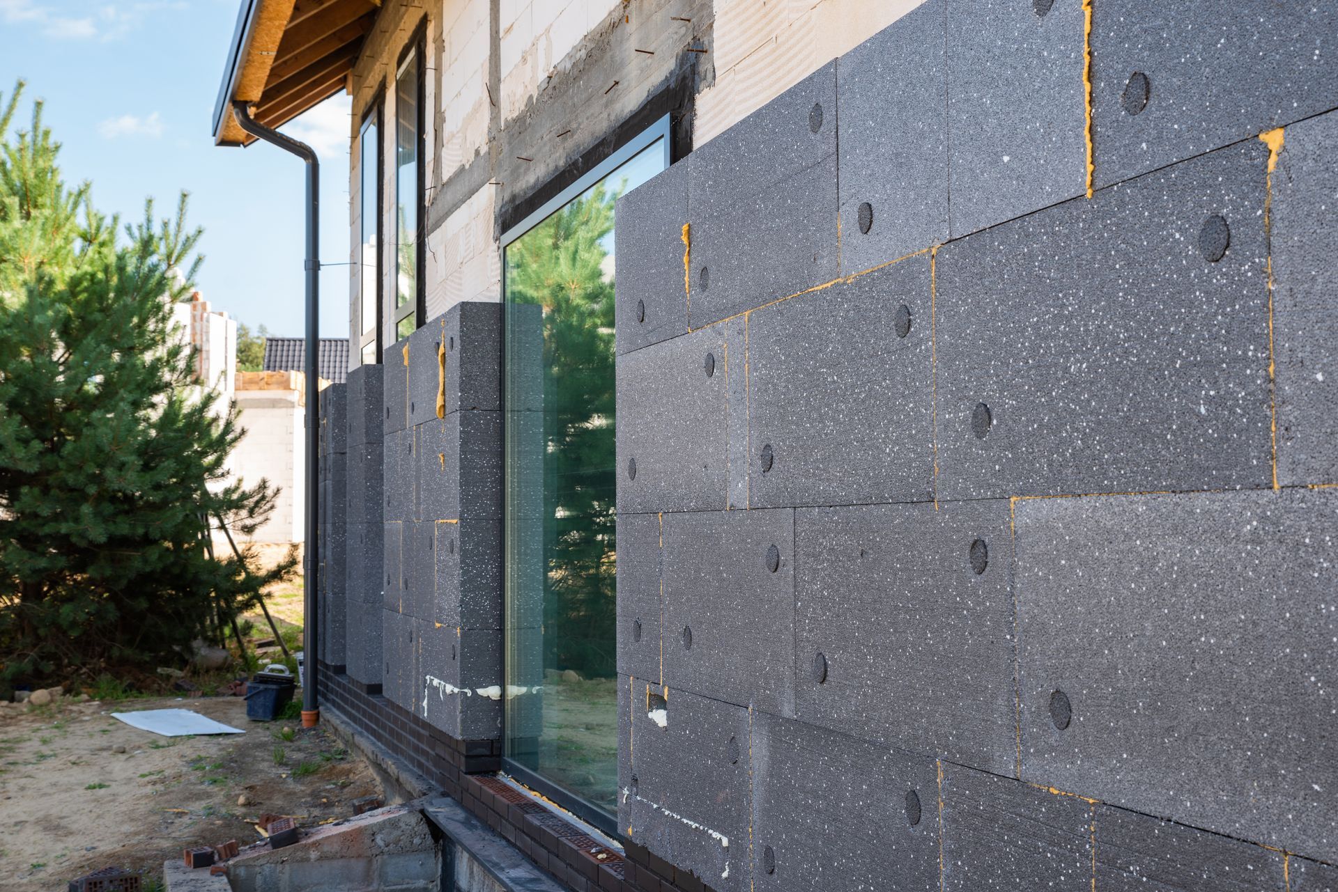 Mur extérieur en construction avec panneaux d'isolation en mousse grise et une grande fenêtre donnant sur une cour bordée d'arbres