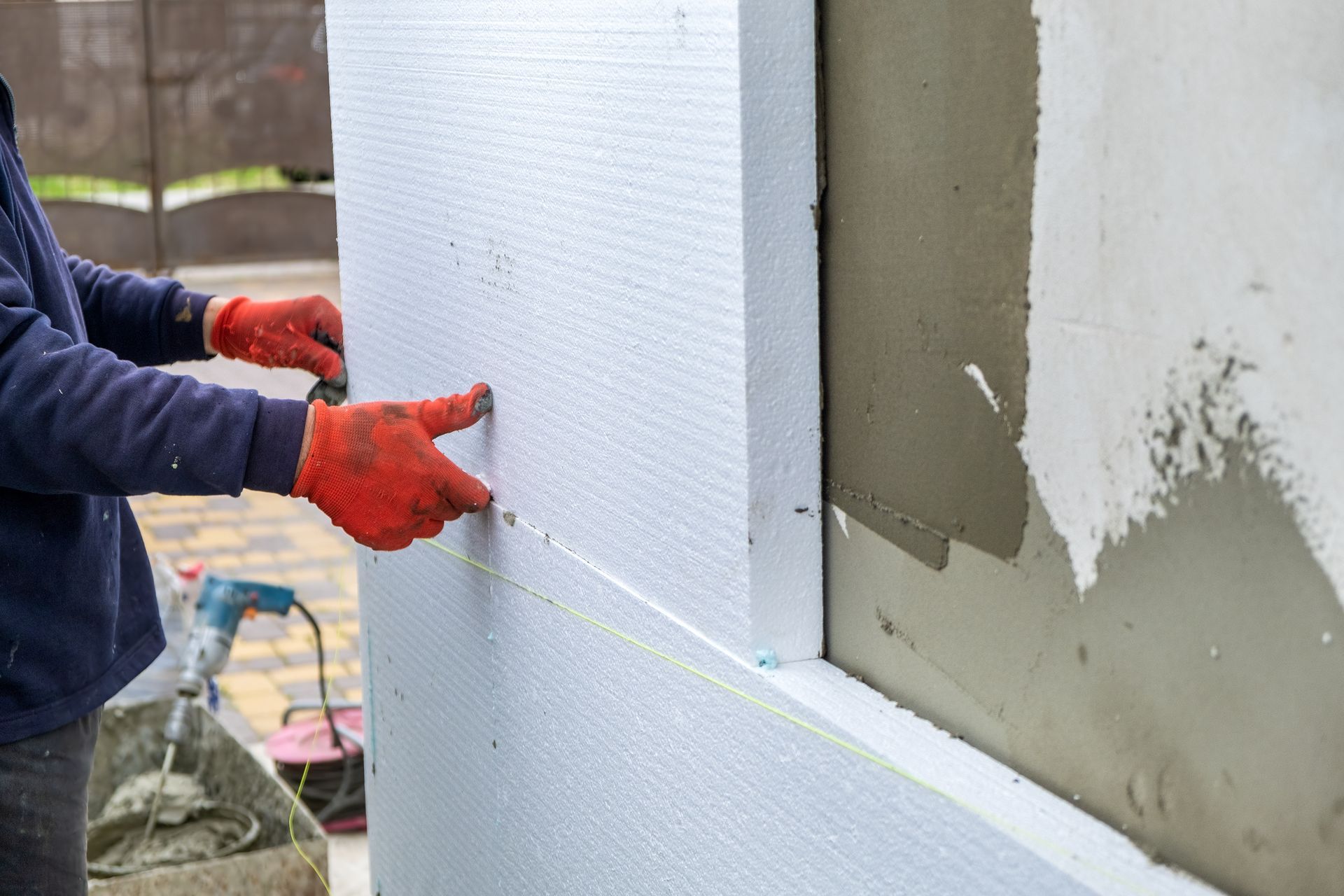Ouvrier installant des panneaux d'isolation en mousse blanche sur un mur extérieur, portant des gants rouges.