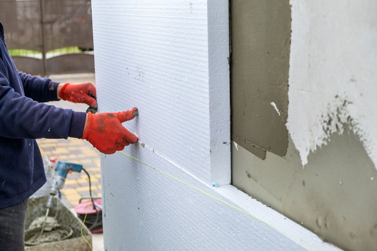 Ouvrier installant des panneaux d'isolation en mousse blanche sur un mur extérieur, portant des gants rouges.