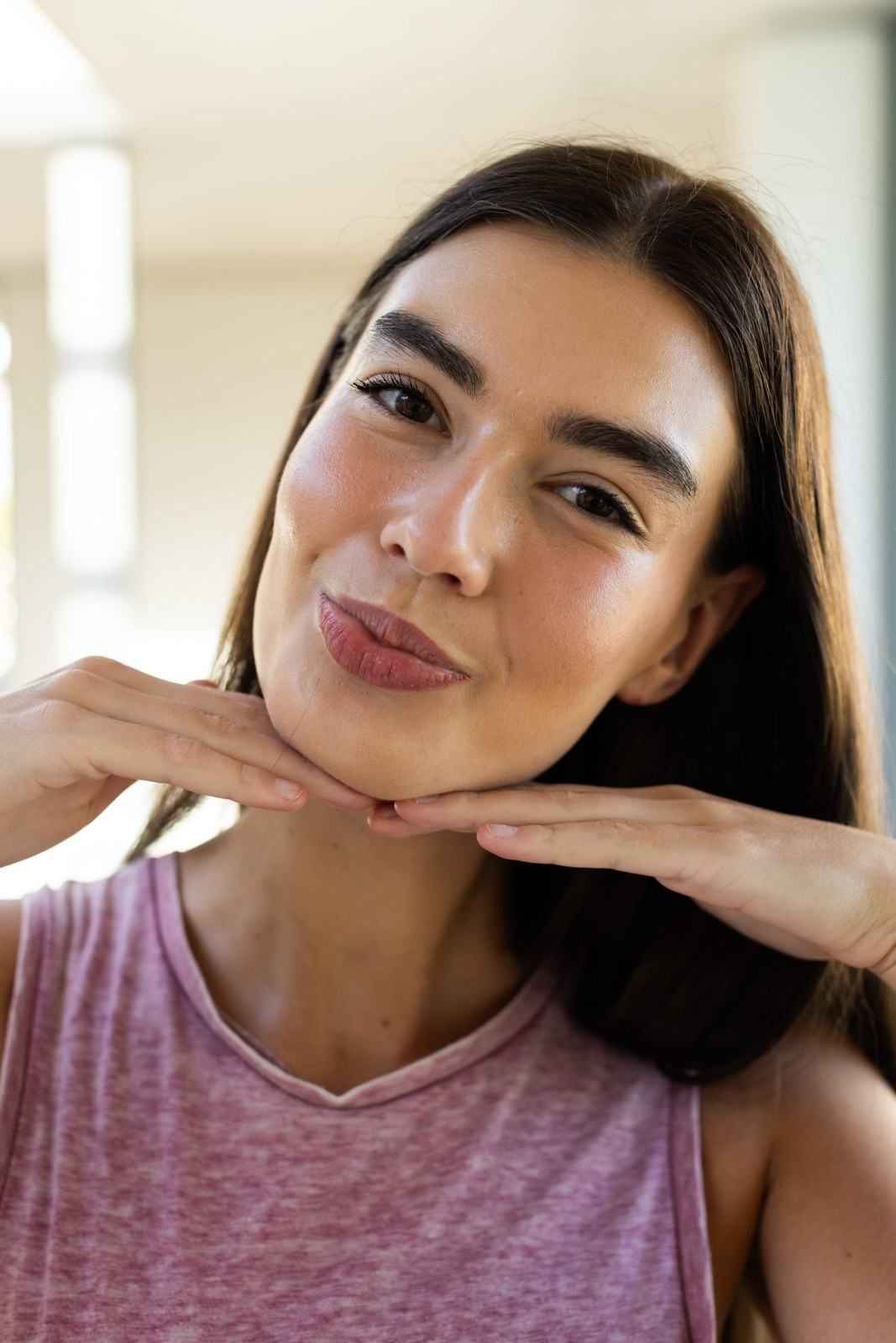 Mujer con un top rosa sin mangas, sonriendo con las manos bajo la barbilla en un espacio interior bien iluminado