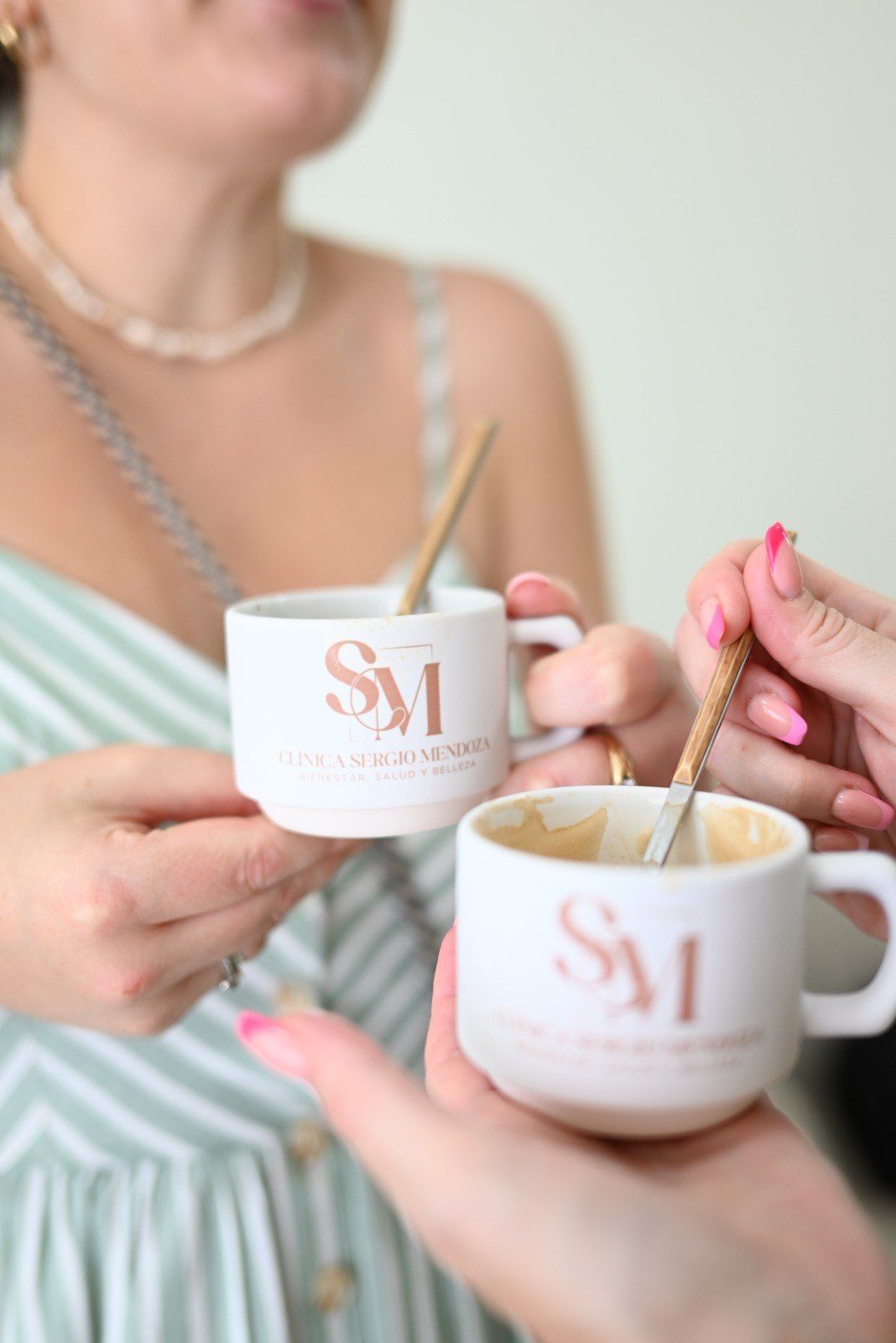 Dos personas sosteniendo tazas blancas con cucharas doradas, una de ellas con un vestido a rayas, en un espacio interior luminoso
