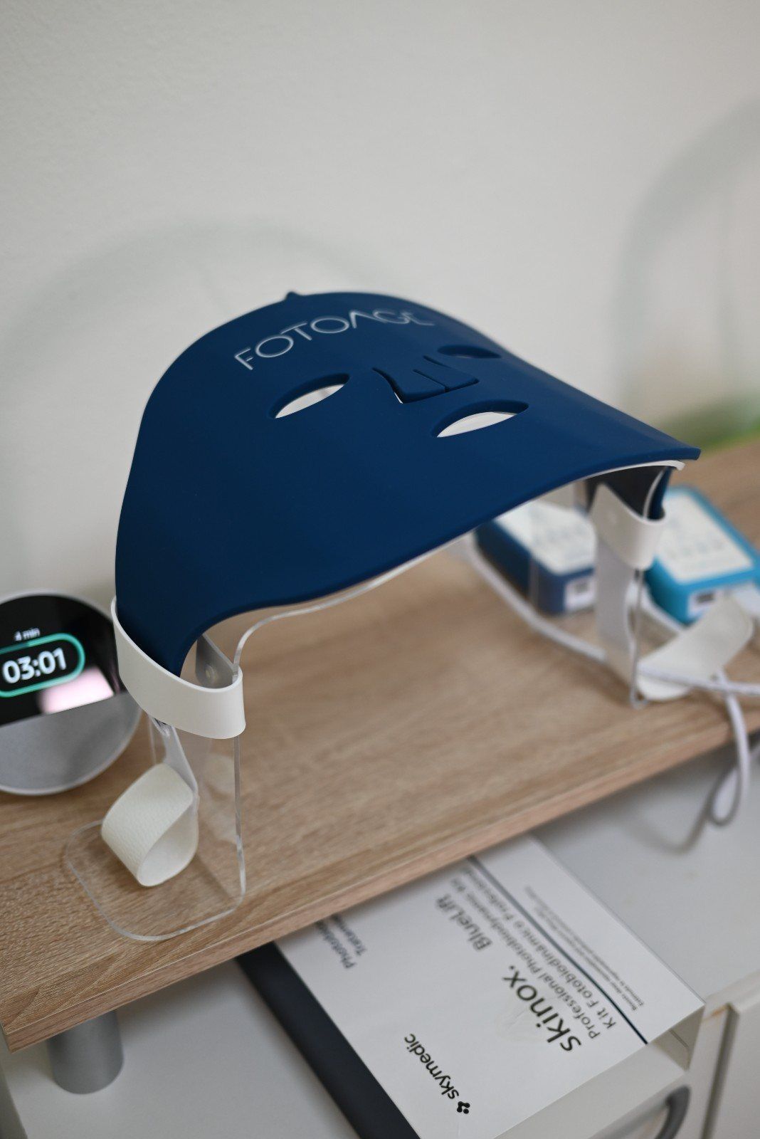 Mascarilla de fototerapia facial azul sobre una estantería de madera, con un pequeño reloj y un cable de carga cerca