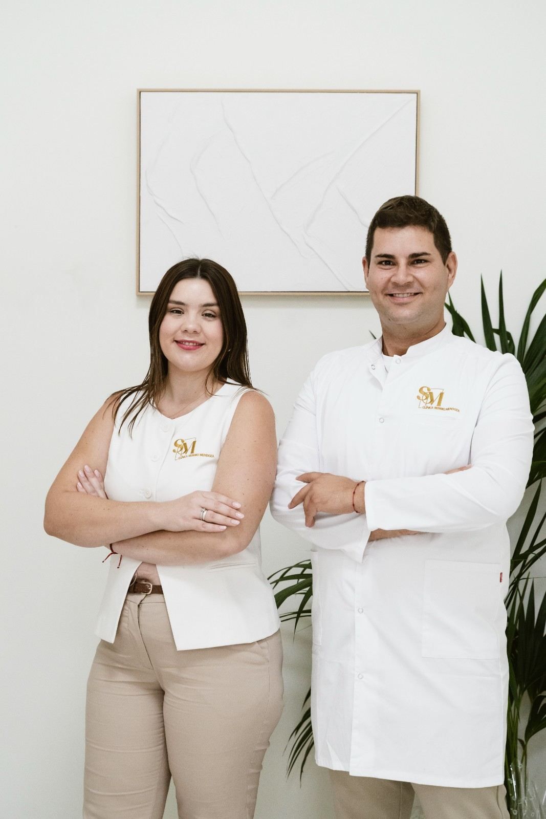 Dos personas con uniformes blancos están de pie con los brazos cruzados en una oficina luminosa.